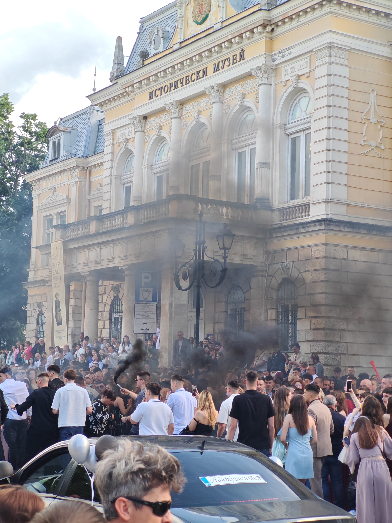 The image shows a crowd of people gathered in front of a building identified by a sign as the "Исторически музей" (Historical Museum) in Ruse, Bulgaria. Black smoke is emanating from the crowd. People are dressed in a mix of casual and formal attire. Some individuals are holding phones, potentially recording the event. Balloons are attached to a car, and a license plate is visible with Cyrillic characters on it. The architecture of the building is characterized by columns and arched windows.