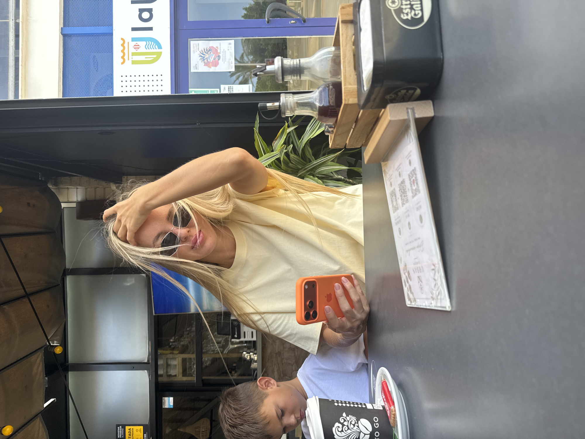 A young woman with blonde hair and sunglasses is holding an orange smartphone and taking a selfie at an outdoor cafe. She is wearing a light yellow t-shirt. A young boy in a white t-shirt is visible in the foreground, seemingly looking down at something on the table.
The setting appears to be a cafe or restaurant with an outdoor seating area. In the background, there is a building with blue and white signage that includes text and a logo. There are also some plants and bottles of what appear to be condiments on a wooden stand. A menu or flyer is placed on the table in front of the woman. The lighting suggests it is daytime, possibly late afternoon, with a mix of natural light and shade. The overall mood seems relaxed and casual.
Visible text includes "la Vila Joiosa" and "LA VILA JOIOSA" on the signage in the background, along with "MOSTAR" and "EXITADA A POLICIA" on a yellow sticker on a wooden structure in the lower left of the image. The coffee cup in the foreground has text that includes "TO GO". The woman's phone case is bright orange.