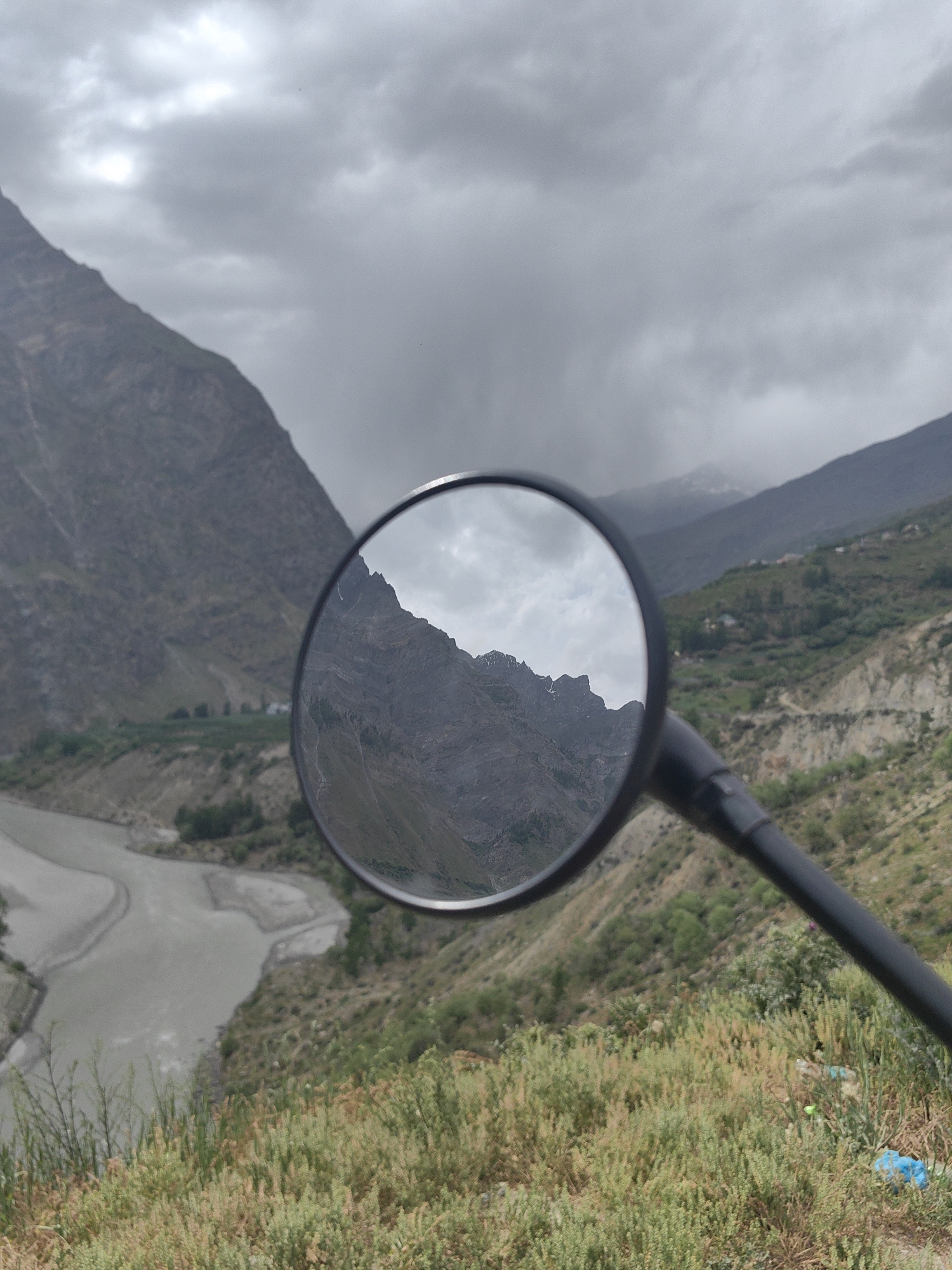 A motorcycle rearview mirror reflects a mountainous landscape with a river and cloudy skies. The mirror is mounted on a black handlebar. The landscape in the reflection shows rugged, gray mountains with patches of green vegetation and a winding river below. The foreground of the image, outside the mirror's reflection, features dry grass and some greenery. The sky is overcast with clouds. The scene appears to be in Tandi, India, given the mountainous terrain and river, characteristic of the region. There are no people or specific actions depicted.