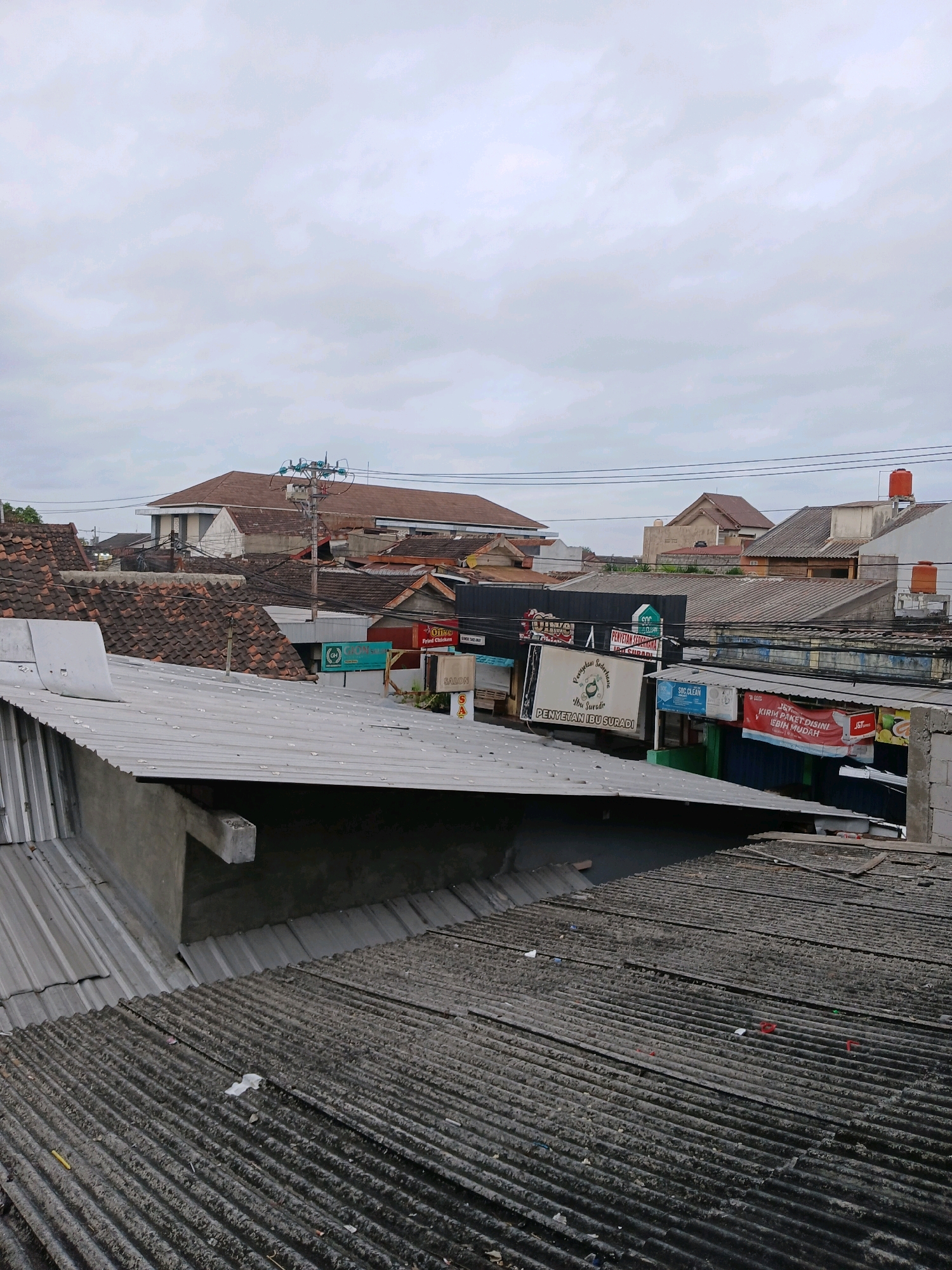 The image captures a view of rooftops in Depok, Indonesia, under an overcast sky. The foreground is dominated by corrugated metal roofing, some of which appears weathered and dusty. In the mid-ground, a variety of buildings with tiled roofs are visible, along with some commercial signs. These signs include "J&T Express", "SGC CLEAN", and an establishment named "PENYETAN IBUSURADI". Further in the background, larger buildings and more roofing are seen, along with power lines and a utility pole. There are no people or discernible activity in the scene. The overall impression is of a densely built urban or suburban area.