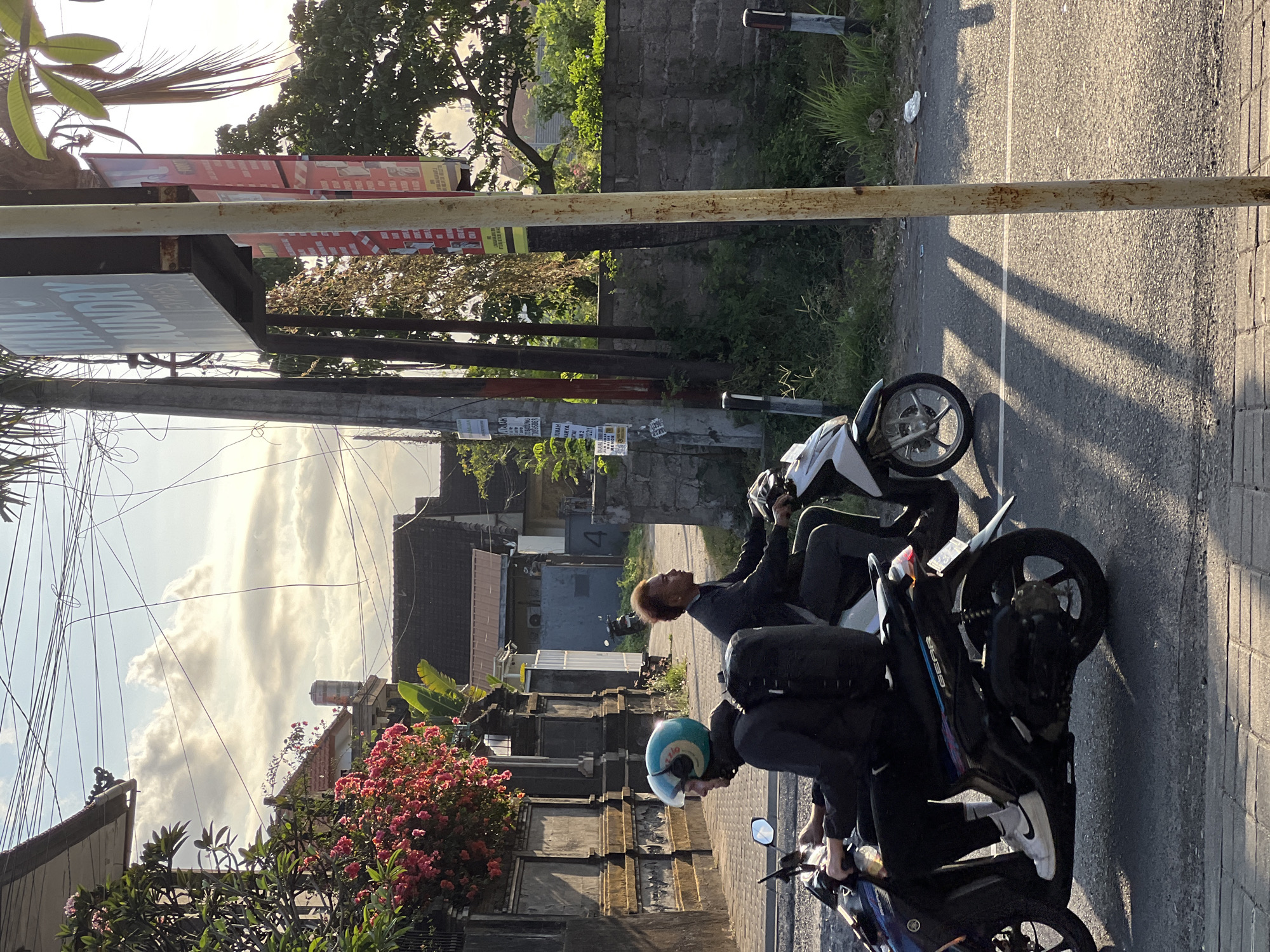 In Denpasar, Indonesia, at sunset, two individuals are riding motorbikes on a paved street. The person in the foreground, wearing a blue helmet and a black jacket, rides a blue motorbike. The second individual, with reddish-brown hair, rides a white motorbike alongside. Behind them, houses with tiled roofs and an ornate wall suggest a residential or mixed-use area. A sign for "MANA LAUNDRY EXPRESS" is visible on a pole. The environment is illuminated by the warm glow of the setting sun, casting long shadows across the road.
