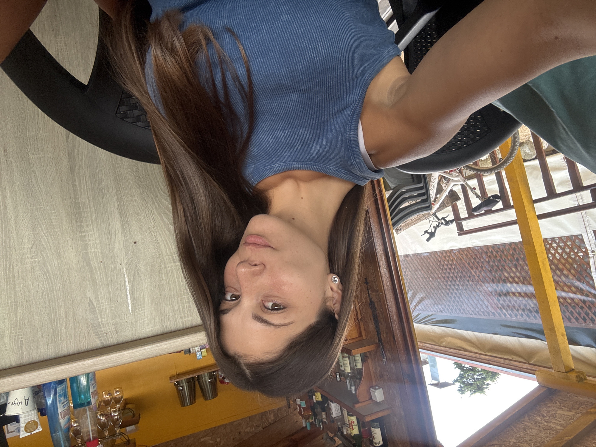 A young woman with long brown hair is taking a selfie outdoors. She is wearing a blue tank top and is seated in a black chair. Behind her, a bicycle is visible, and further in the background, there's a structure with shelves stocked with bottles. The scene appears to be in a casual outdoor setting, possibly a cafe or bar, in Sozopol, Bulgaria. The woman's face is visible, and she is looking towards the camera. Her right arm is extended, holding the phone for the selfie. The overall lighting suggests it is daytime.