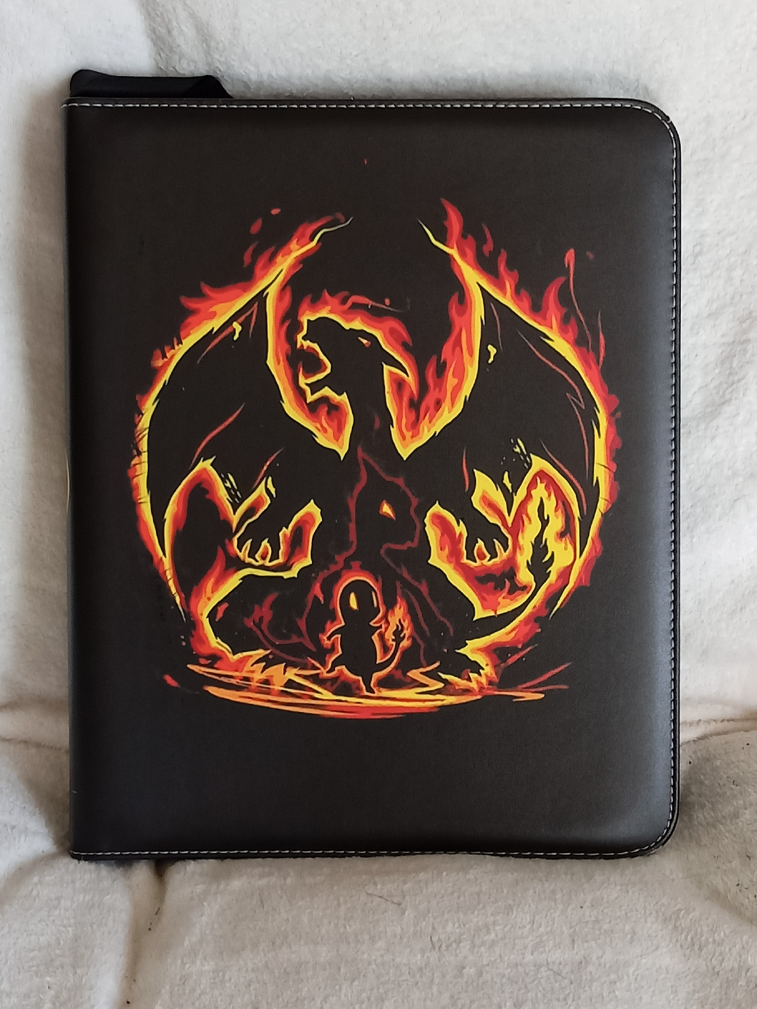 A black binder with a printed image of Charizard and Charmander from Pokémon surrounded by flames is lying flat on a white, fuzzy surface. The binder has a dark grey tab sticking out of the top left corner and white stitching along the edges. The image features a silhouette of a large Charizard with its wings spread, and a smaller Charmander in front of it, both depicted as being engulfed in or made of fire, with shades of orange, yellow, and red. The binder is positioned on a textured fabric that appears to be a blanket or rug. The lighting suggests an indoor environment, with no indication of the city of Amarillo, United States of America.