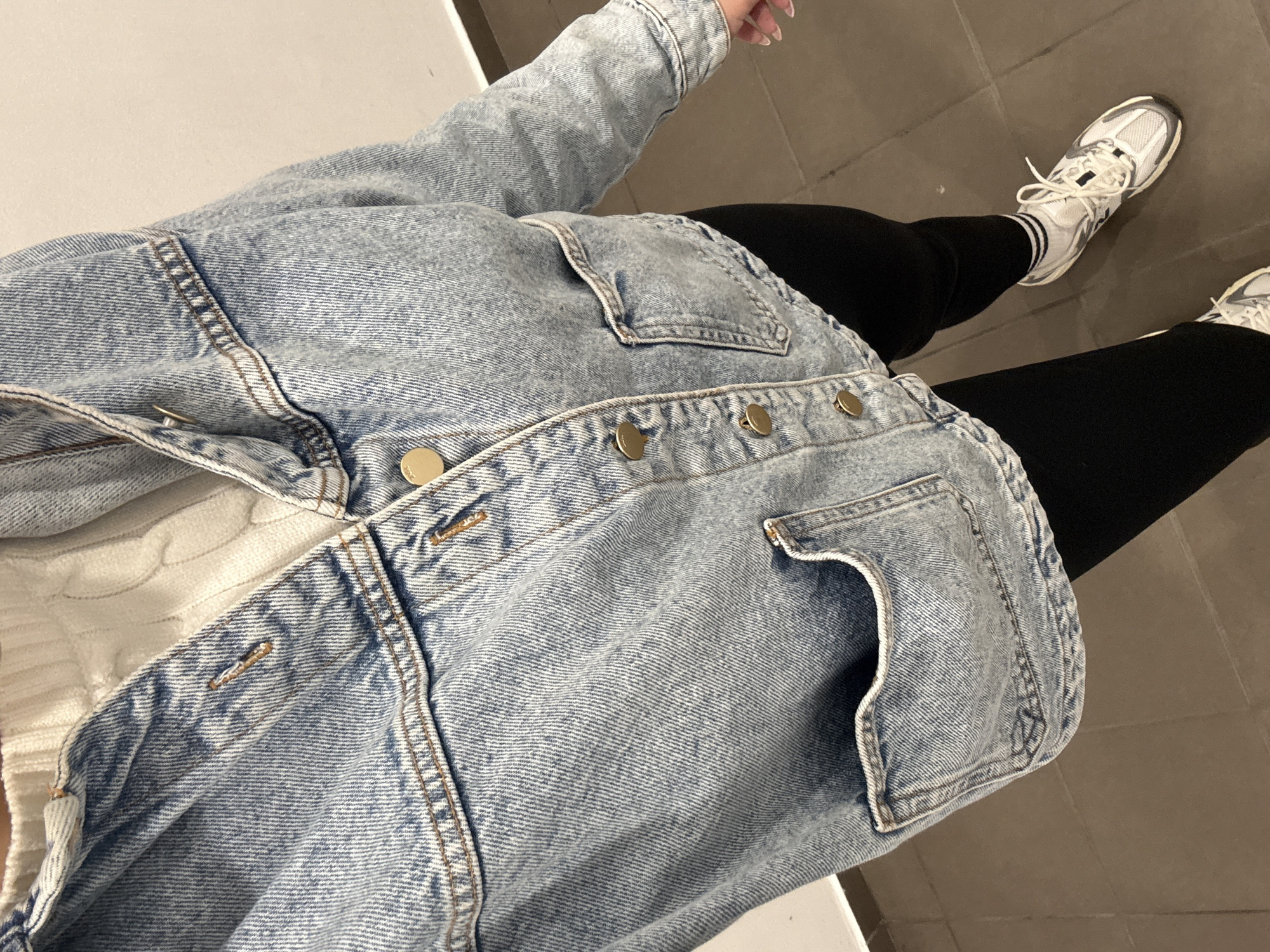 A person wearing a light blue denim jacket, a white cable-knit sweater, black leggings, and white sneakers is standing on a gray tiled floor. The jacket is buttoned with gold buttons and has front pockets. The leggings are form-fitting and extend down to the ankles. The sneakers have white laces and a gray sole. The location is unknown and cannot be determined as Plovdiv, Bulgaria.