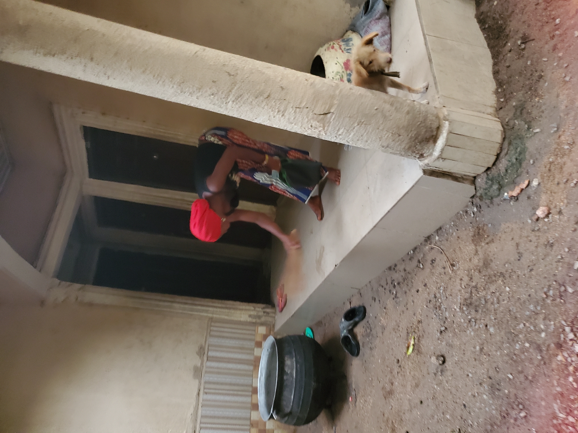The image shows a scene in Mararaba, Nigeria. A woman wearing a red head covering and a patterned dress is on a porch, sweeping dirt into a cloud of dust. Near her is a large, dark-colored cooking pot with a metal rim. A pair of black sandals lie nearby.
On the porch, behind the woman, is a light-brown dog with a stick in its mouth. The porch is supported by a thick, weathered column. The ground surrounding the porch appears to be dirt and gravel. The building next to the porch features a wall with what appears to be a barred window.