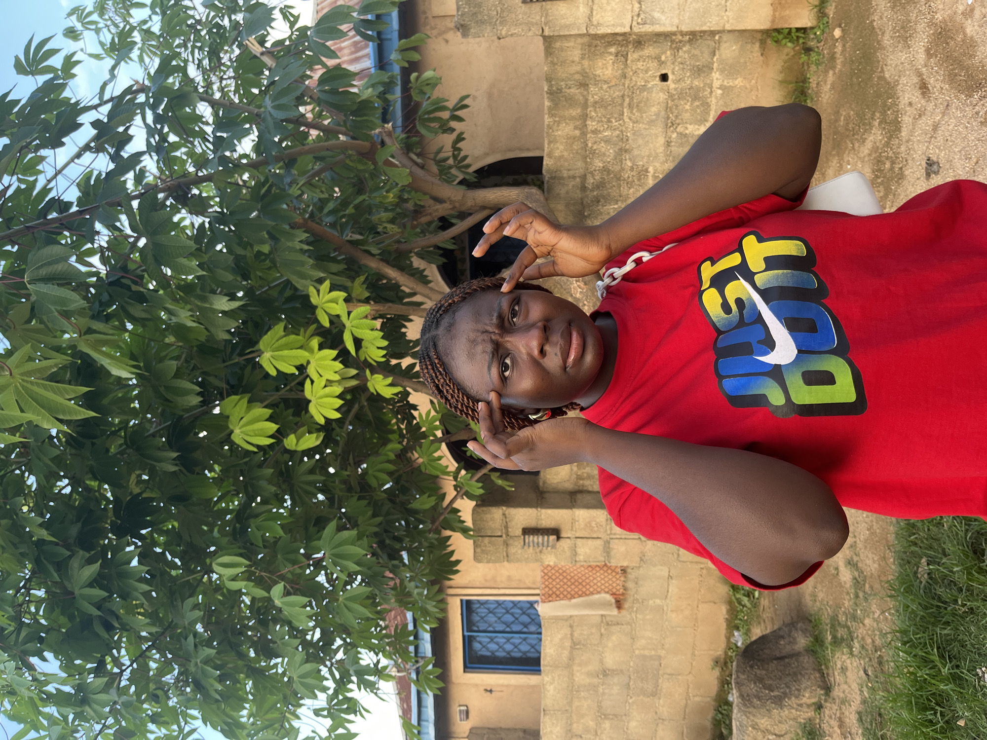 The media file shows a portrait of a young Black woman posing for a photo. She is wearing a red t-shirt with the word "JUST" in bold, colorful letters and a Nike swoosh. Her hair is braided, and she has a thoughtful or confused expression on her face, with her fingers near her temples. She is standing outdoors, with a large green tree with broad leaves to her left. Behind her, there is a building constructed of light-colored bricks, with a blue-barred window visible. The ground is a mixture of dirt and possibly gravel. The lighting suggests it is daytime, possibly late afternoon, with soft shadows. The overall setting appears to be a casual, possibly rural or semi-urban environment.