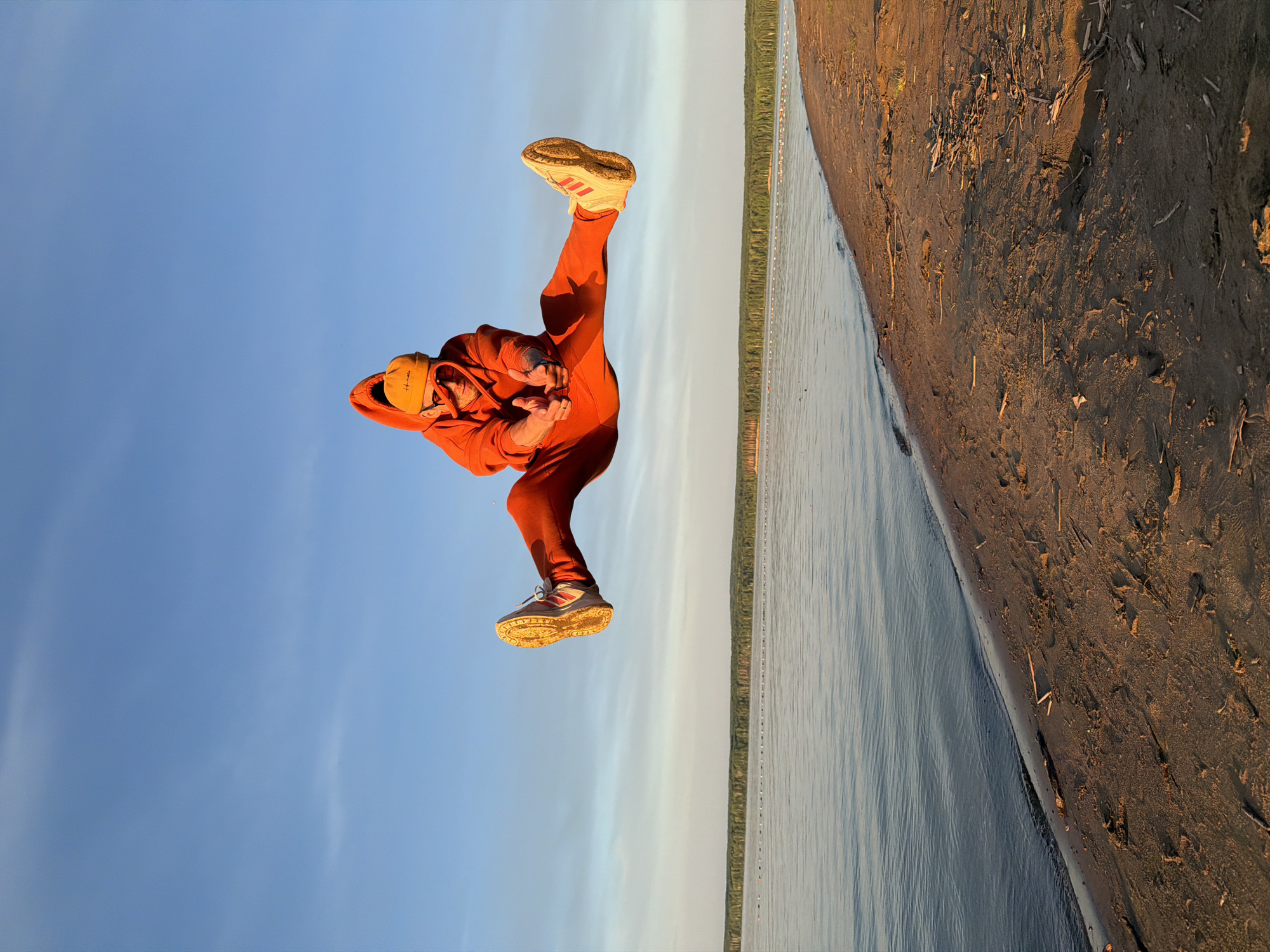 The image captures an individual in a dynamic, athletic pose at a riverside or lakeside location, possibly near Chaikovsky, Russia. The primary subject is a person, likely male, dressed in a vibrant orange hooded tracksuit and an orange or yellow beanie. They are wearing light-colored athletic shoes with red stripes, which appear scuffed or dirty on the soles, suggesting activity. The person is mid-action, with one leg lifted high and bent at the knee, and arms bent, held close to the body with hands seemingly clasped in front.
The scene is set outdoors under a clear, bright blue sky. To the left of the image is a calm body of water, possibly a river or lake, with subtle ripples on its surface. Extending across the water into the distance is a line of small buoys or markers, suggesting a designated area or path. To the right is a sandy or muddy bank, rich in earthy brown and golden tones, with scattered debris like twigs and small pieces of wood. A distant treeline of dark green foliage is visible across the water.
The lighting indicates either late afternoon or early morning, with warm, golden sunlight illuminating the bank and the person, casting subtle shadows. The person's pose suggests an energetic activity, possibly a jump, dance move, or martial arts stance, conveying focus and dynamism. On the beanie, some light-colored, indistinct text is visible, possibly a brand name.