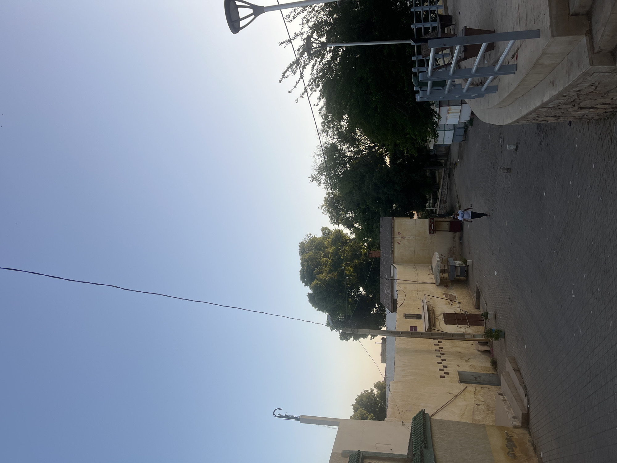 The image depicts a street scene in El Hajeb, Morocco. The perspective is from an elevated viewpoint looking down a paved street lined with aged, light-colored buildings. A solitary individual, wearing a white t-shirt and dark pants, walks away from the camera down the street. To the right of the street, a raised walkway with a metal railing and benches is visible, overlooking a lower level with trees and more buildings. A modern streetlamp stands prominently on this raised area. The sky is a clear, pale blue, suggesting it is either early morning or late afternoon. The architecture is characteristic of Moroccan urban environments, with textured walls and traditional building elements. The street itself is paved with interlocking gray bricks, and there are a few potted plants and small market stalls along the sides of the buildings.
