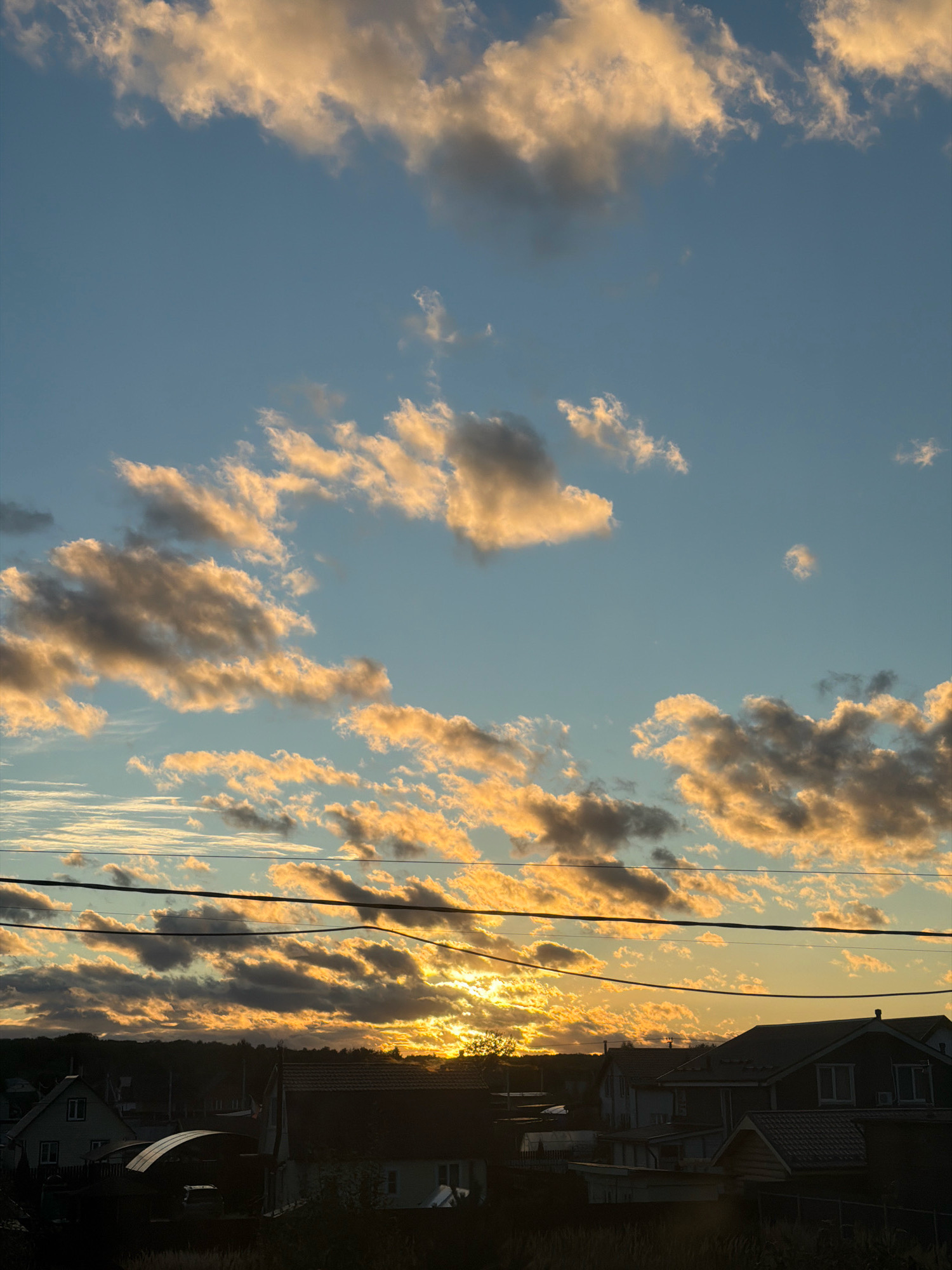 This is a photograph of a sunset over a residential area in Stupino, Russia. The sky is filled with clouds that are illuminated by the setting sun, casting warm golden and orange hues. Below the sky, power lines are visible, stretching horizontally across the frame. The lower portion of the image depicts a silhouette of houses and trees against the bright sky. The overall impression is one of a peaceful evening scene.