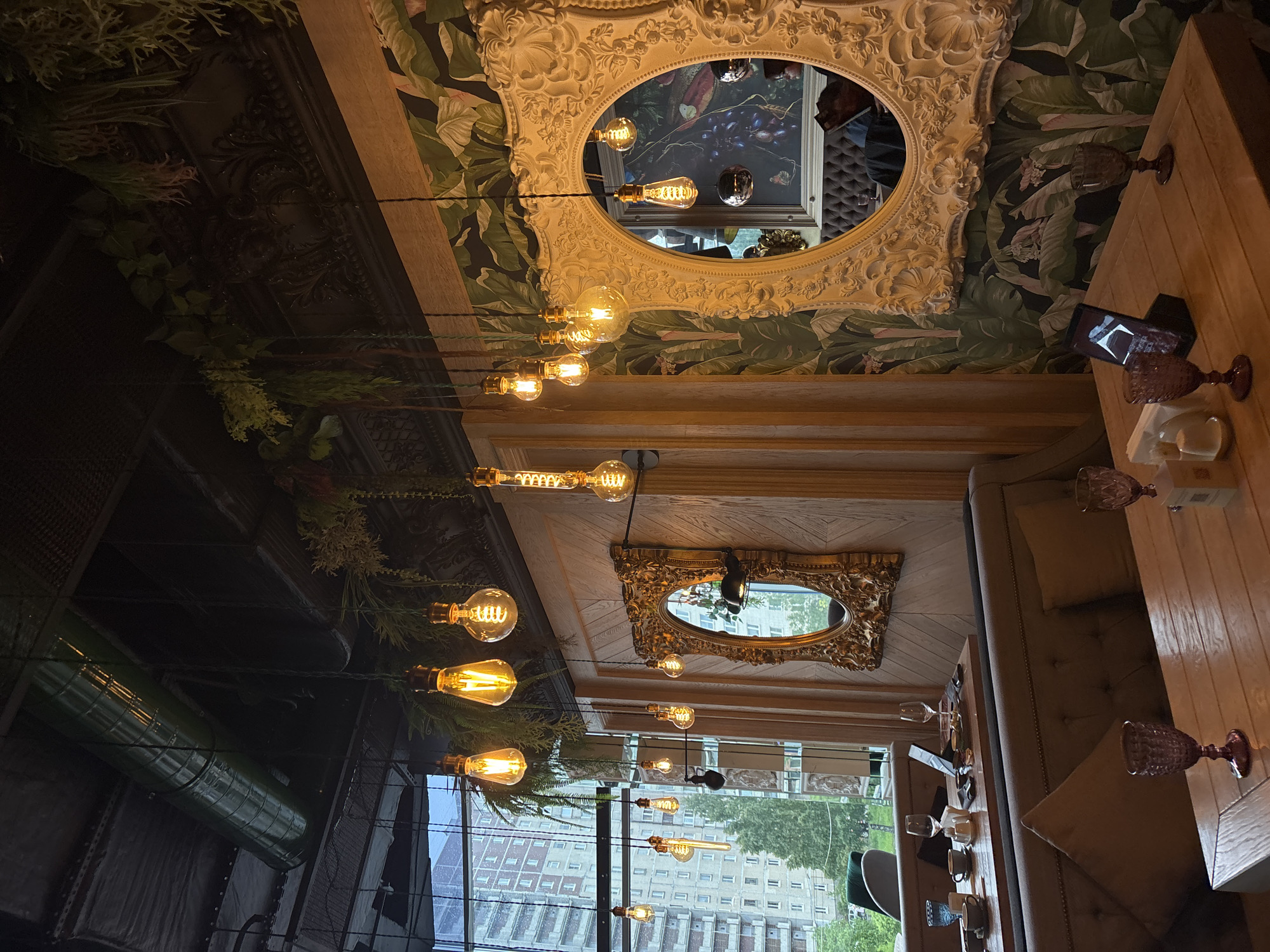The image captures the interior of a likely upscale restaurant in Saint Petersburg, Russia. The vantage point is from below, looking upwards towards the ceiling. The ceiling features ornate floral wallpaper and decorative elements, including two prominent framed mirrors, one oval and one rectangular, both with intricate gold or white detailing. A series of vintage-style Edison light bulbs hang at varying heights from the ceiling, suspended by thin cables.
The scene includes reflections of the interior, visible within the mirrors. To the right, a wooden table setting is visible, complete with glassware, napkins, and what appears to be a mobile phone. A cushioned seating area runs alongside the table. The external world visible through the large window, reveals a view of a tall building in Saint Petersburg.