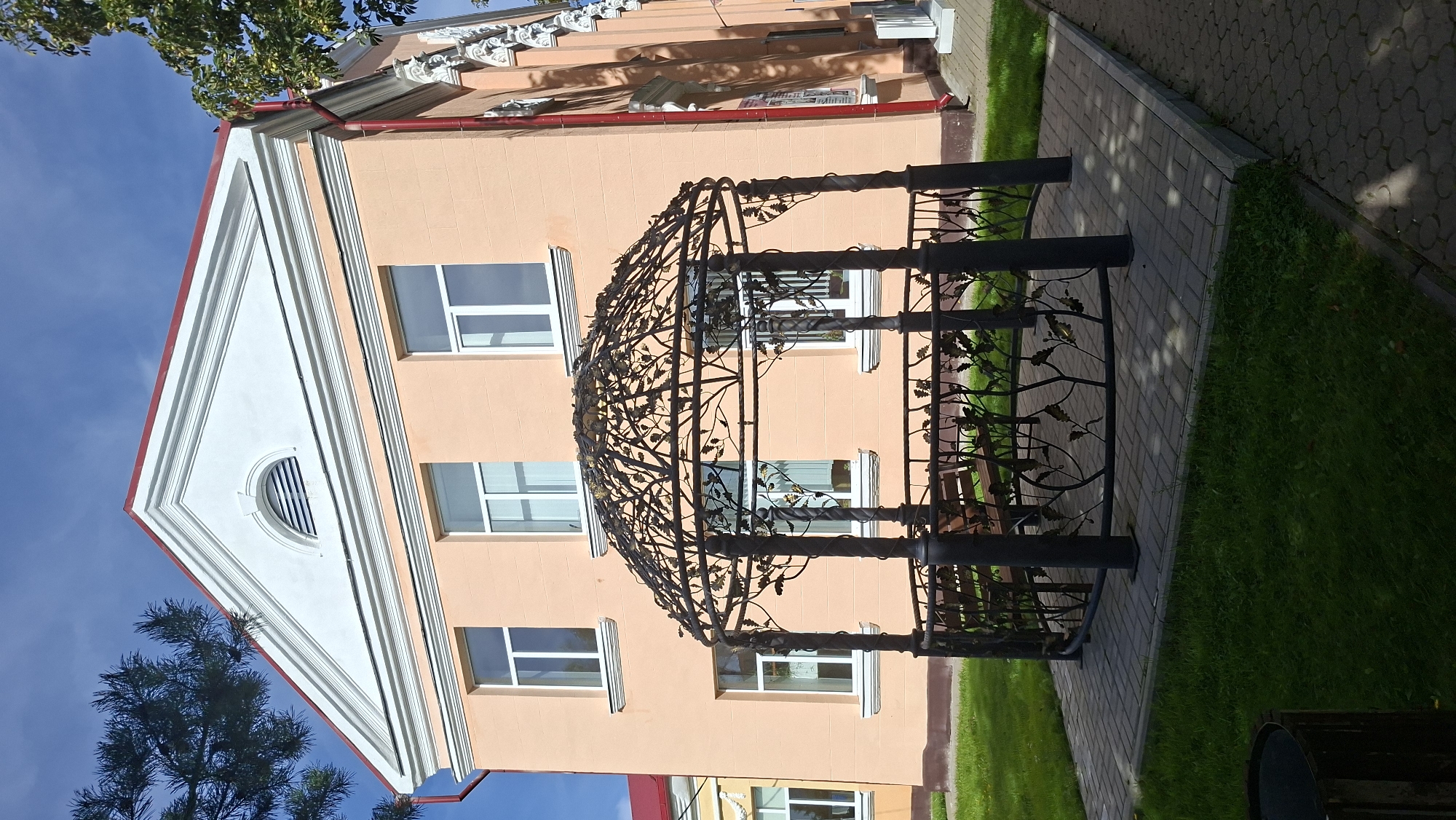 A wrought-iron gazebo with a leafy design stands on a paved area in front of a peach-colored building. The gazebo has dark posts and a decorative roof. A wooden bench is visible inside the gazebo. The building has white-trimmed windows and a white triangular pediment with a semi-circular vent at the top. Red gutters run down the side of the building. The ground around the gazebo is covered in grass and paving stones. The sky is blue with some clouds. This scene is located in the city of Myadzyel, Belarus.