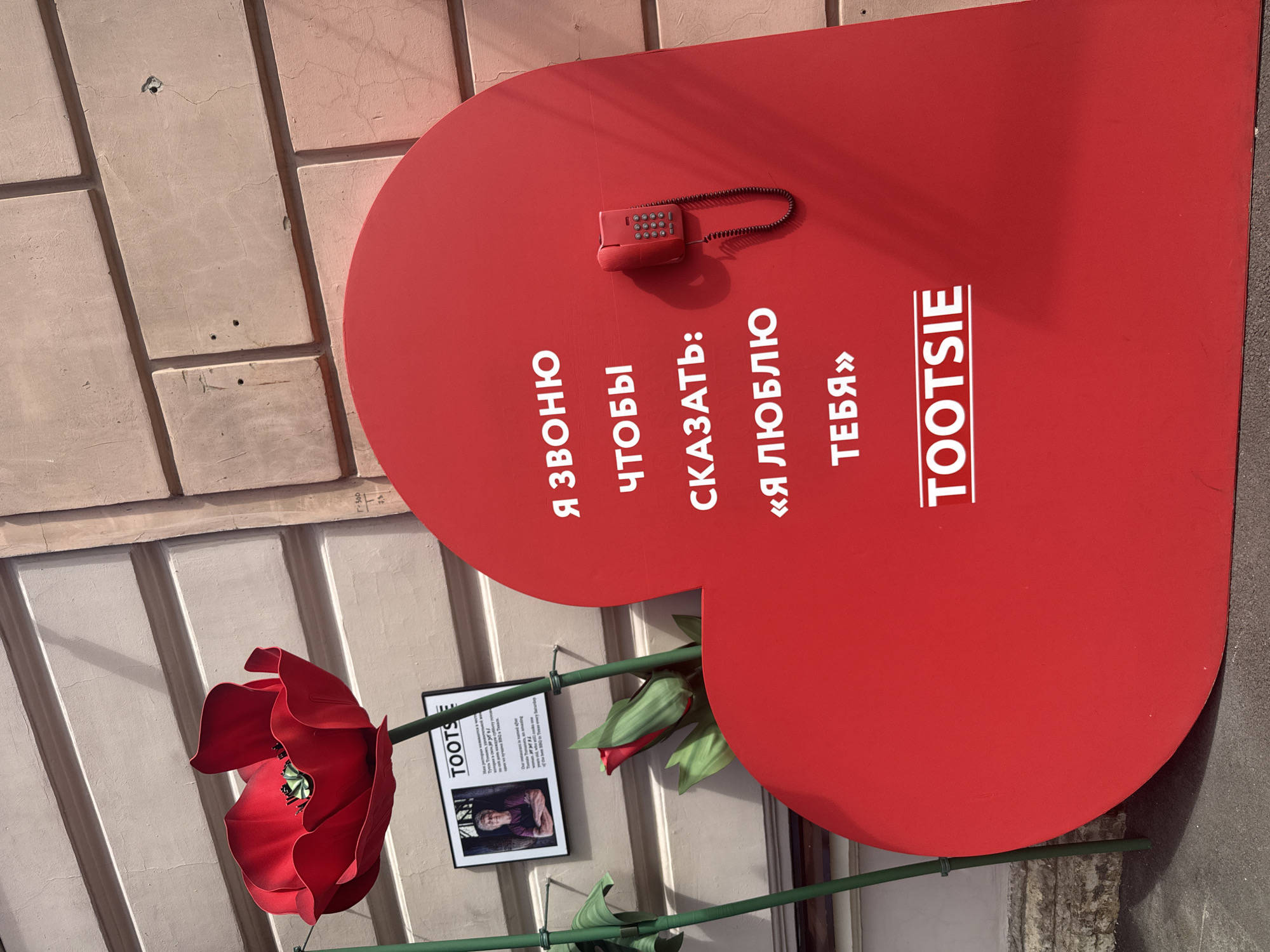 In Saint Petersburg, Russia, a large, bright red heart-shaped installation stands against a light-colored brick wall. The heart features white text in Russian that translates to "I'm calling to say: 'I love you' Tootsie". A vintage red rotary telephone is attached to the surface of the heart. To the left of the heart, a large, artificial red poppy with green stems is positioned, with its bloom reaching upwards. Below the poppy, a framed black and white photograph is affixed to the wall, displaying a woman. The image captures an outdoor urban setting, likely a street or public square, on a bright day with shadows cast by the installation and the poppy.
