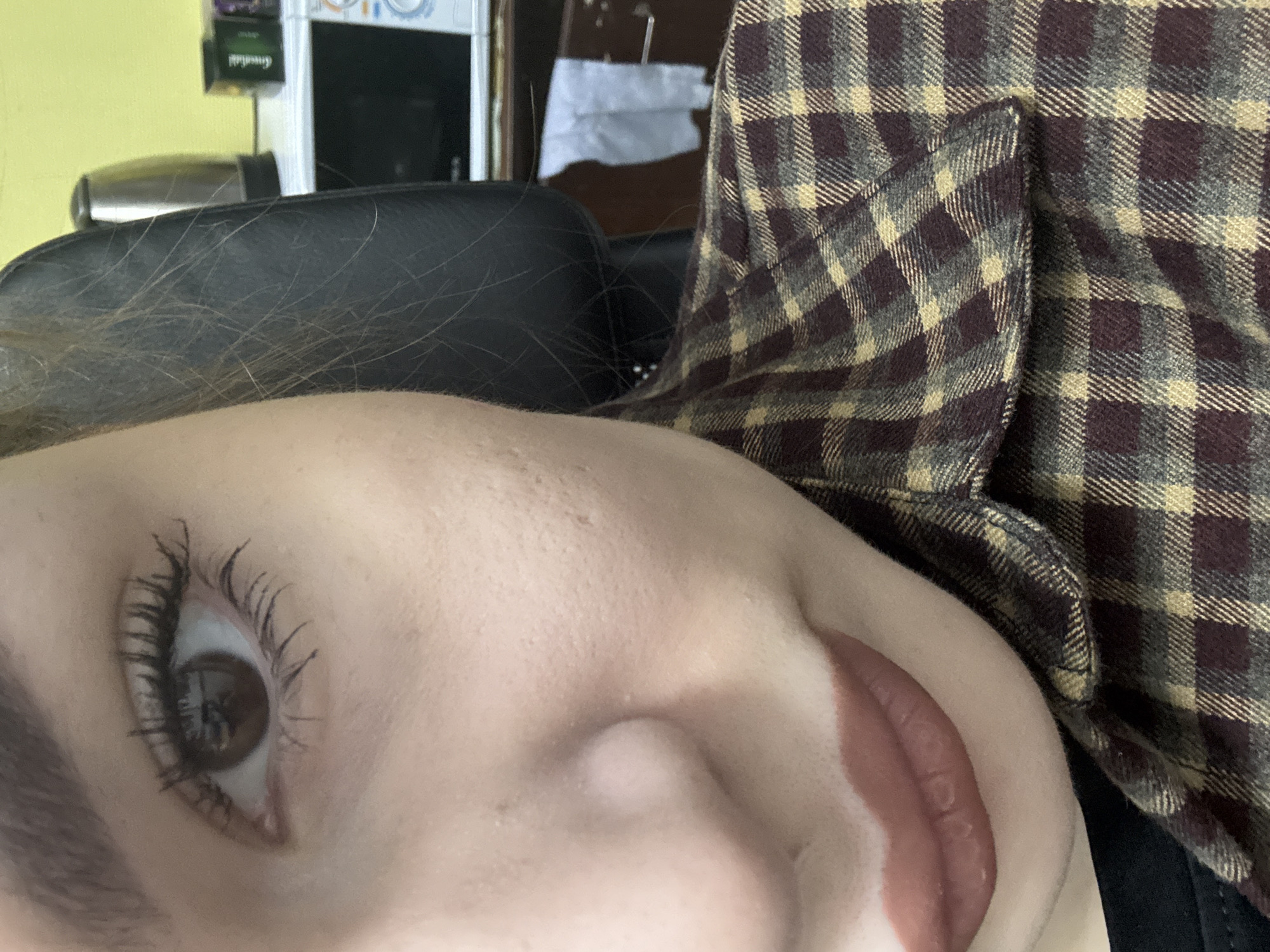 A close-up, low-angle shot focuses on a person's face. The individual has fair skin, an eye with prominent lashes coated in mascara, and lips coated with lipstick. Part of their face rests on a black, padded surface. The individual is wearing a flannel shirt in a brown and tan checkered pattern. In the background, there is a hint of a yellow wall, a blurred object that appears to be a hair dryer, and a cabinet or shelf with various items. There is no indication of the location being Chaikovsky, Russia or any information to geolocate the picture.