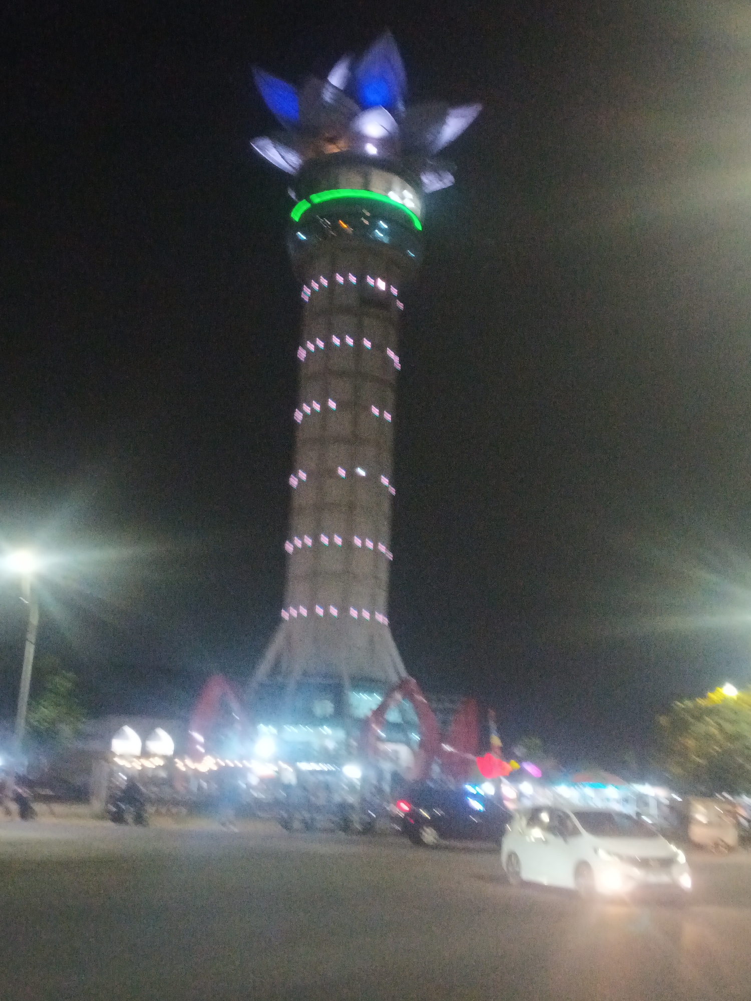 This is a nighttime photograph of the Tugu Pahlawan (Heroes Monument) in Purwokerto, Indonesia. The monument is a tall, slender tower illuminated with pink lights in horizontal bands and a green band just below the top. The very top of the tower is adorned with blue and white structures that resemble petals. In the foreground, a white car with its headlights on is driving on a paved road. The background shows a street scene with other vehicles, including motorcycles, and various lights from buildings and possibly market stalls. The overall impression is of a vibrant, well-lit urban environment at night.