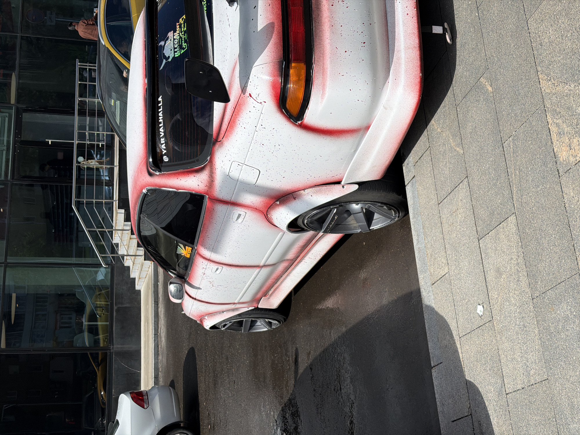 A white car with red spray paint accents is parked on asphalt next to a sidewalk. The car is positioned at an angle, with its rear facing the sidewalk. A portion of a building with reflective windows and a stairway is visible in the background, along with other parked vehicles. The scene appears to be outdoors during daylight.