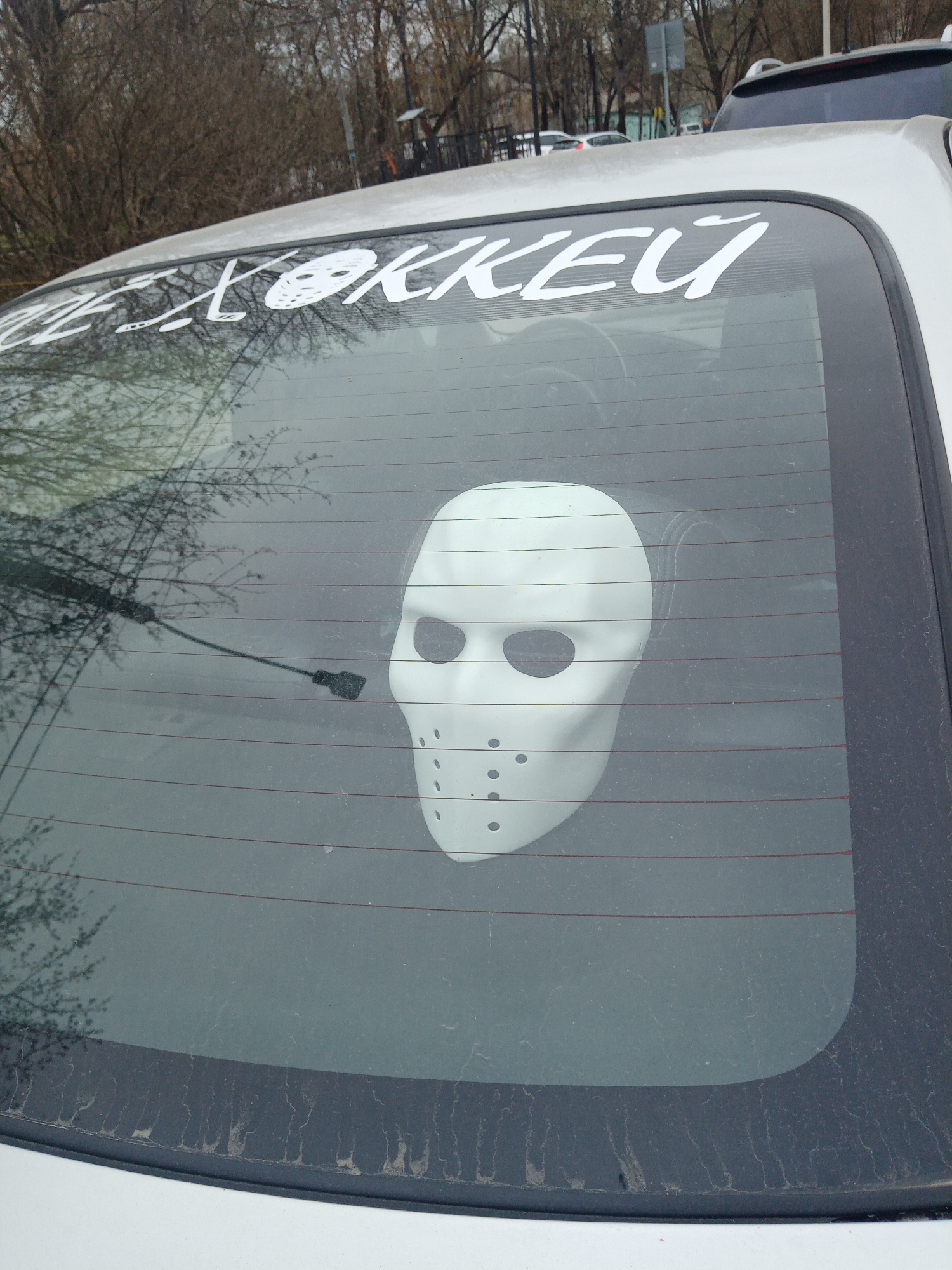 The image shows the rear window of a light-colored car. A white hockey mask decal is affixed to the center of the rear window. A white sticker with text in a Cyrillic alphabet, possibly reading "ХОККЕЙ", is placed at the top of the window. The car is parked outdoors, with trees and other vehicles visible in the background. The rear window is visibly dirty. The location is identified as Elektrostal, Russia.