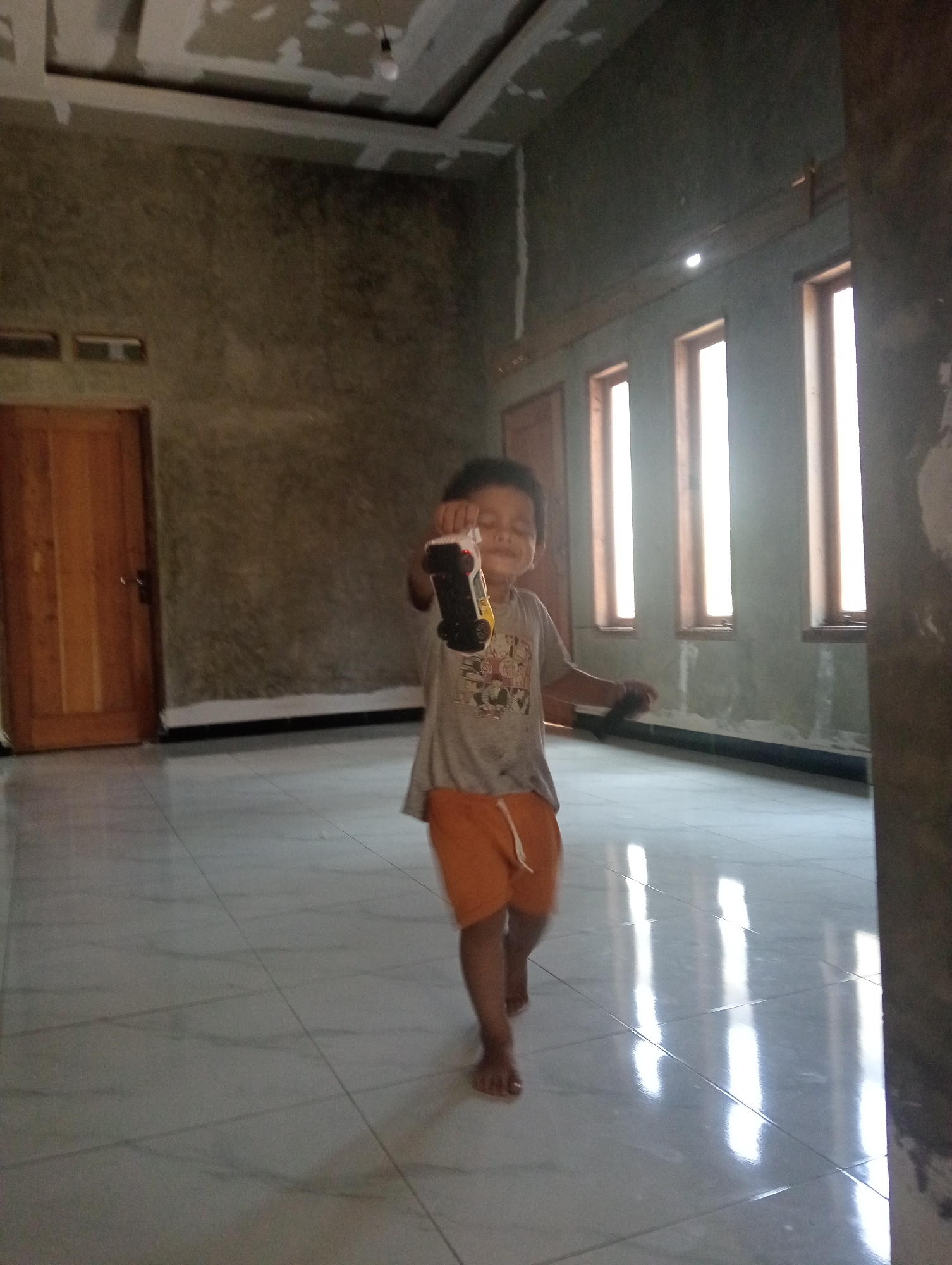 An Indonesian child, appearing to be a boy between the ages of 4 and 7, is captured mid-stride in what seems to be an unfinished residential interior. He is wearing a grey t-shirt with a graphic print and orange shorts. The child is holding up a toy, possibly a model car, with his right hand, and his body is angled slightly towards the viewer. His eyes are closed or squinting, and he has a slight smile, suggesting he is engaged in play or perhaps posing. The environment is characterized by bare, textured walls, a partially finished ceiling with exposed drywall and wiring, and large, vertical windows that allow bright light to stream in, creating reflections on the tiled floor. A wooden door is visible on the left side of the frame. The scene suggests a home under construction or renovation in Muara, Indonesia.