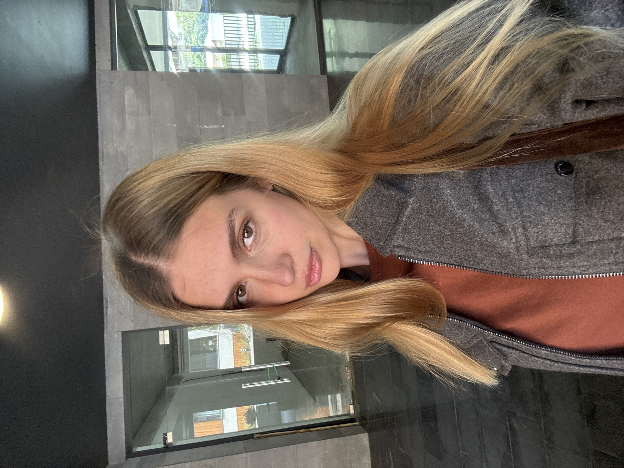 A fair-skinned woman with long, blonde hair is shown in a close-up, angled portrait. She has brown eyes, light eyebrows, and slightly parted lips. She is wearing a brown shirt with a gray wool jacket. The setting appears to be an indoor space with large glass windows and gray tiled walls. Reflected in the glass is a view of a building with white and brown exterior walls, suggesting an urban environment.