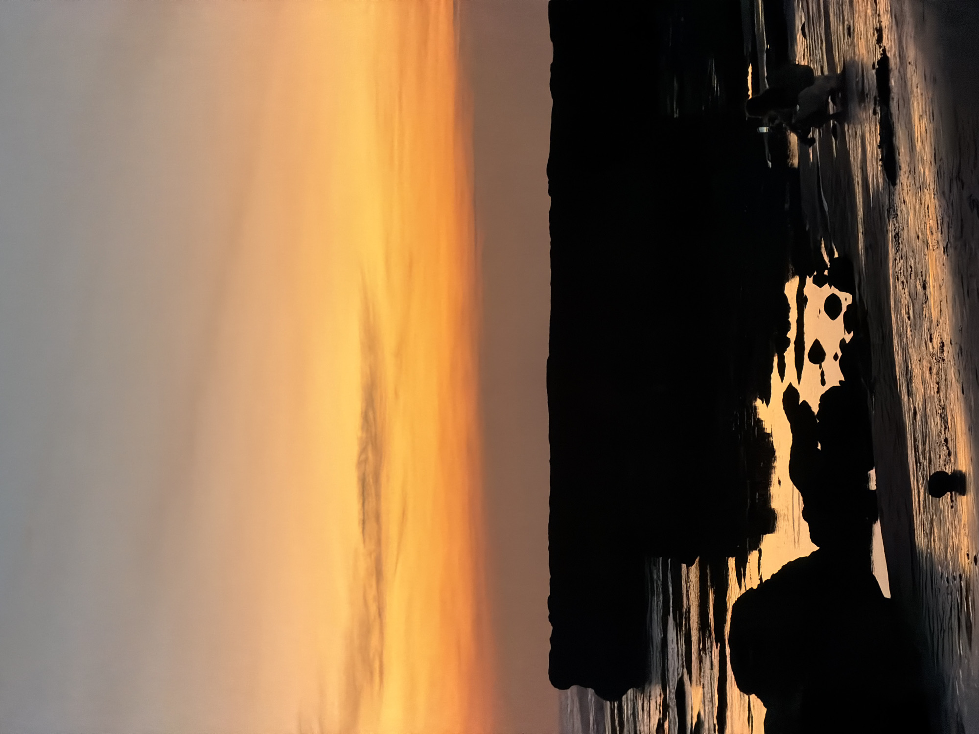 A full shot captures a serene sunset on the coast of North Kuta, Indonesia. The sky above is a gradient of soft orange and pale blue, with wispy clouds tinged with yellow. In the foreground, the dark silhouettes of large rocks and boulders dot the wet sand, reflecting the warm glow of the sunset. The gentle waves of the ocean create a shimmering pattern on the beach. A lone figure, silhouetted and crouched on the sand, is engrossed in taking a picture with their phone, their posture suggesting a quiet moment of appreciation for the natural beauty. The overall scene evokes a sense of tranquility and the peaceful end of a day at the beach.