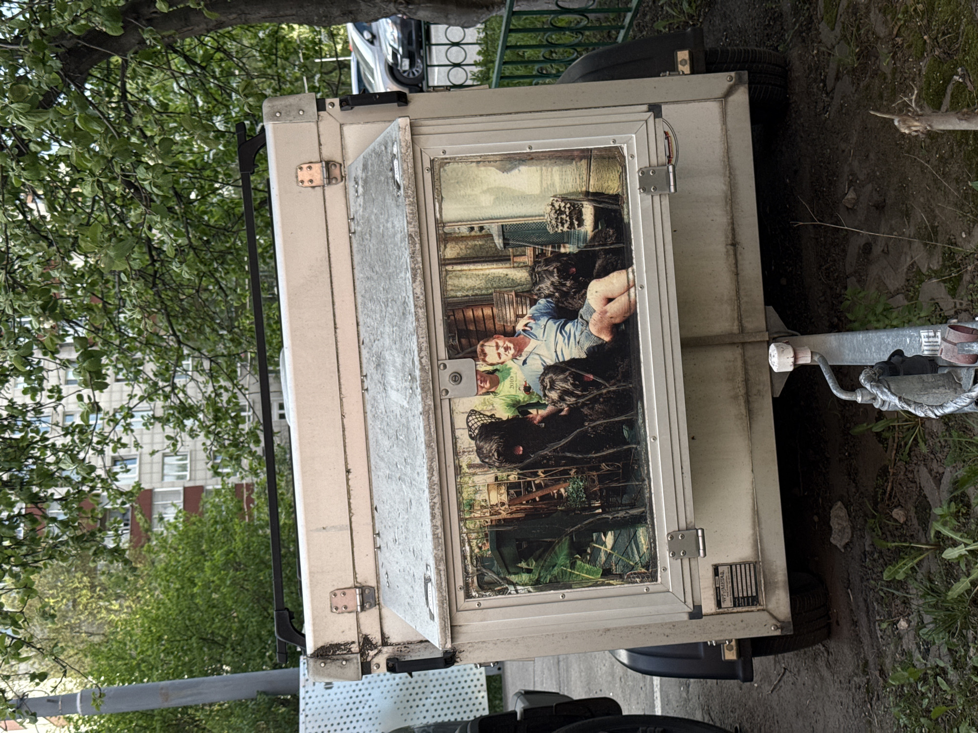The image shows a small, boxy trailer parked on what appears to be a patch of ground next to a road or sidewalk. The trailer has a light gray or off-white exterior with metallic trim and hinges. A large print is attached to the side of the trailer, featuring a photograph of a man lying among ferns and what appears to be a large dog. The photo is somewhat obscured. The trailer is connected to a tow hitch. A tree is visible behind the trailer on the left side of the frame, and a building with many windows is visible in the background. A section of a metal fence can be seen above the trailer. The location is identified as Moscow, Russia.