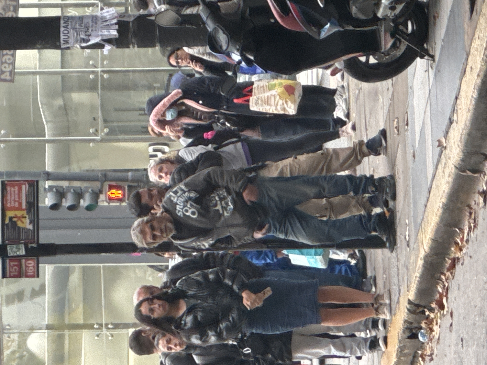 A daytime street scene in Buenos Aires, Argentina features a cluster of people standing on the sidewalk near a bus stop with signage displaying numbers "151" and "160," and a traffic light showing a red signal. Several individuals are visible, some with shopping bags, and a few are walking. A motorcycle is parked nearby on the sidewalk. A building is in the background with advertising posters on the windows.