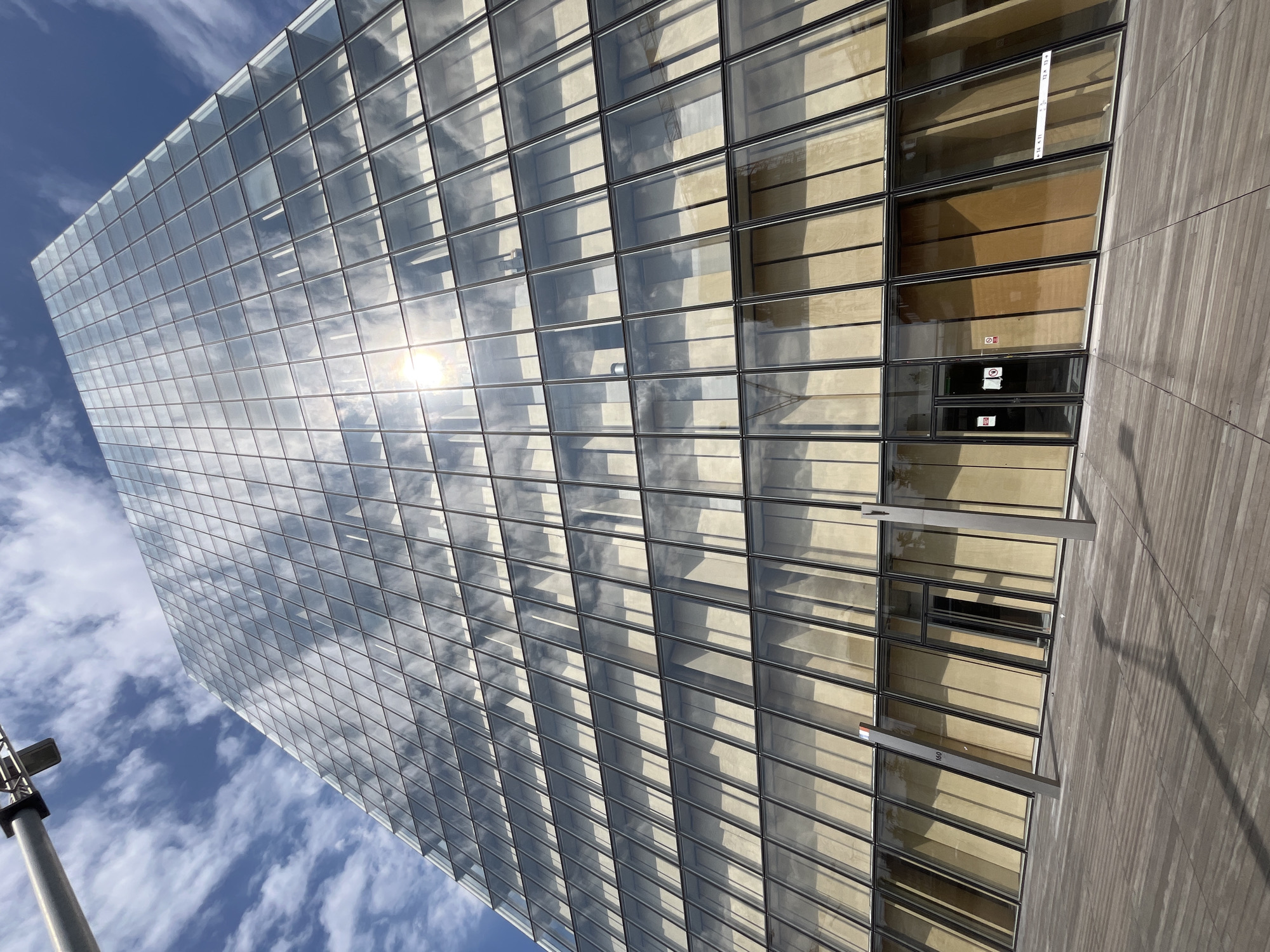 This media file is a photograph of a modern building with a glass facade. The building is tall and its exterior is composed of many rectangular glass panes framed by dark metal. The glass reflects the sky and clouds, creating a dynamic and shimmering effect. The sun is visible in the reflection, suggesting it is daytime.
To the right of the glass facade, a section of the building is covered in a material that appears to be wood or a wood-like composite, with a vertical grain. This wooden section also has a grid-like pattern, similar to the glass facade. A horizontal metal beam extends from the glass section towards the wooden section, creating a sharp line of shadow.
The lower right portion of the image shows a wooden walkway or deck with a textured surface. A tall, grey pole with a light fixture at the top is visible on the left side of the image, extending upwards.
There is some visible text on the building's glass facade. On one of the glass panels, a white sticker with black text appears to have numbers and possibly other characters, such as "16 11" and "12 12". Another small rectangular sign with red and white markings is visible on a lower glass panel, possibly indicating a hazard or warning. A number "160" is visible on a label attached to a horizontal beam.
The overall impression is one of architectural modernity and a bright, clear day. The angle of the shot, looking upwards, emphasizes the height and scale of the building. The activity, if any, is not discernible from the image, but the scene suggests an urban or commercial setting.