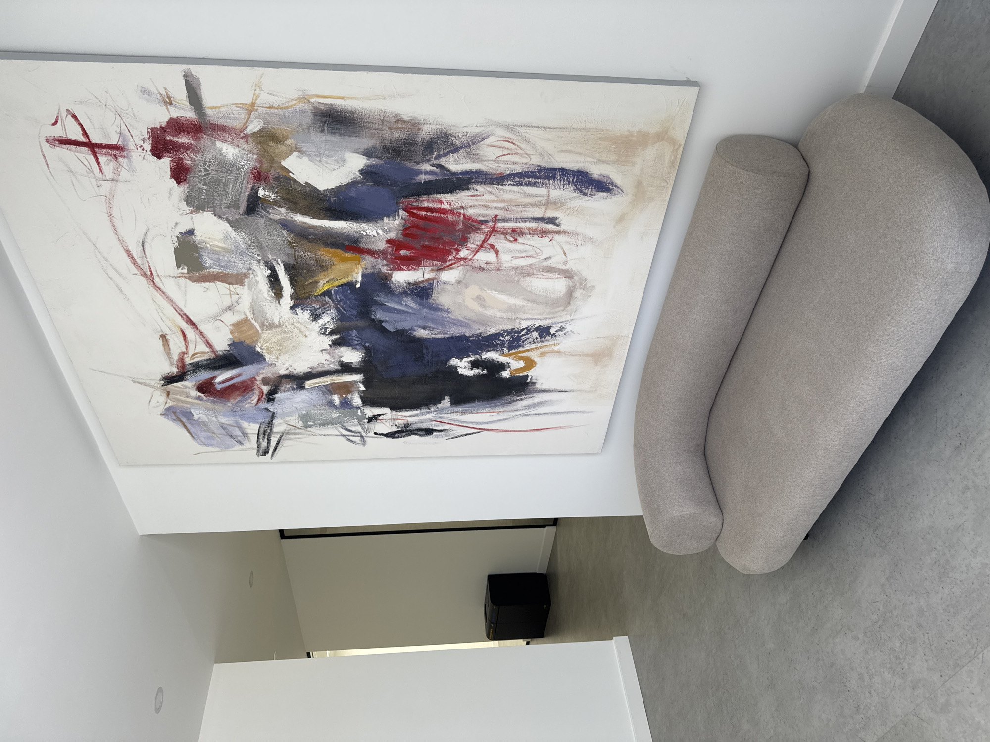 A contemporary interior shot from an elevated perspective shows an abstract painting on a white wall, adjacent to a light gray rounded couch. The painting is composed of red, blue, black, white, and gray brushstrokes. Below the painting, a doorway with a black frame leads to a space with a black cube-shaped object. The floor is gray, and the walls are white. The location is possibly Torres Vedras, Portugal, though this is not explicitly verifiable from the image. No people are visible.