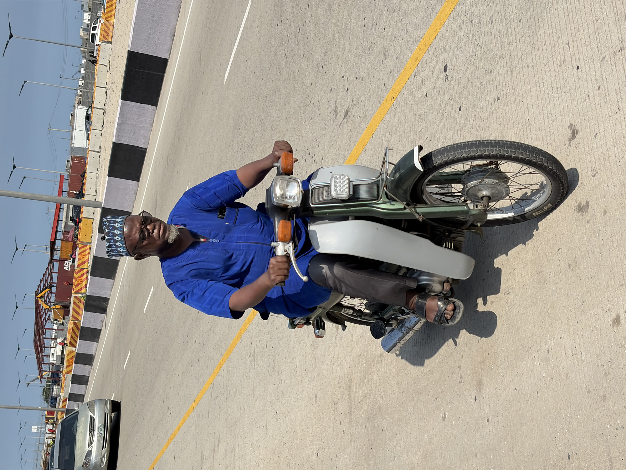 A man with a gray beard and wearing a blue patterned cap and blue shirt is riding a moped on a bridge. He is facing the viewer and has his hands on the handlebars. He is wearing dark pants and sandals. The bridge has a yellow line and a black and gray barrier to the left. In the background, there are power lines and infrastructure. A silver car is also visible. The location is Alaguntan, Nigeria.