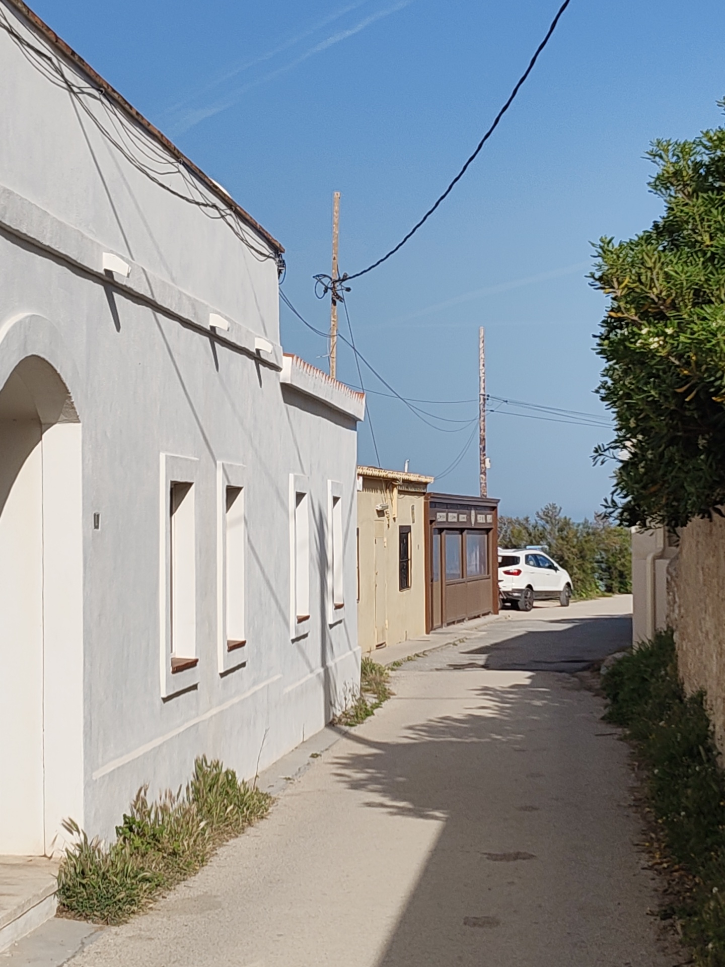 A narrow asphalt road, flanked by buildings, stretches into the distance in Rotes, Spain. On the left, a white building with a light gray façade, marked by three window openings, rises. The bases of its walls are lined with small patches of green vegetation. To the right of this building is a tan structure with an addition of dark brown siding and windows. In the background, a white SUV is parked on the road. Power lines stretch across the sky, connecting wooden utility poles. The sky is clear and blue. A stone wall overgrown with greenery borders the right side of the road, partially obscuring the view. Shadow patterns from unseen elements are cast across the road.