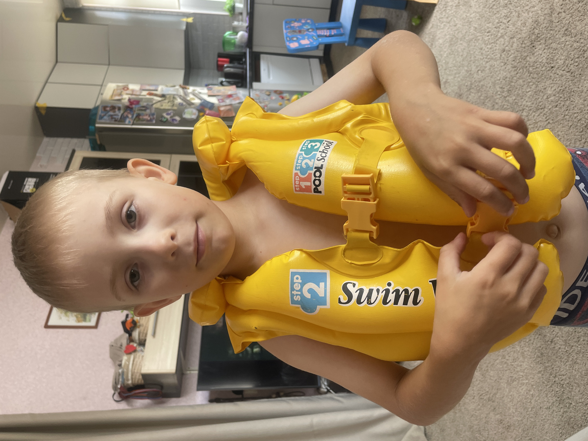 The image shows a young boy wearing a bright yellow swim vest. The vest has a "Step 1 2 3 POOL SCHOOL" graphic on one side and "STEP 2 Swim" on the other, indicating it is designed for learning to swim. The boy's hands are near the clasp of the vest. He is looking towards the camera with a neutral expression. The background suggests an indoor setting, possibly a home, with kitchen cabinets visible. The lighting is indoor lighting, and there are no clear indicators of the time of day or weather. The boy appears to be preparing to swim or has just finished swimming.