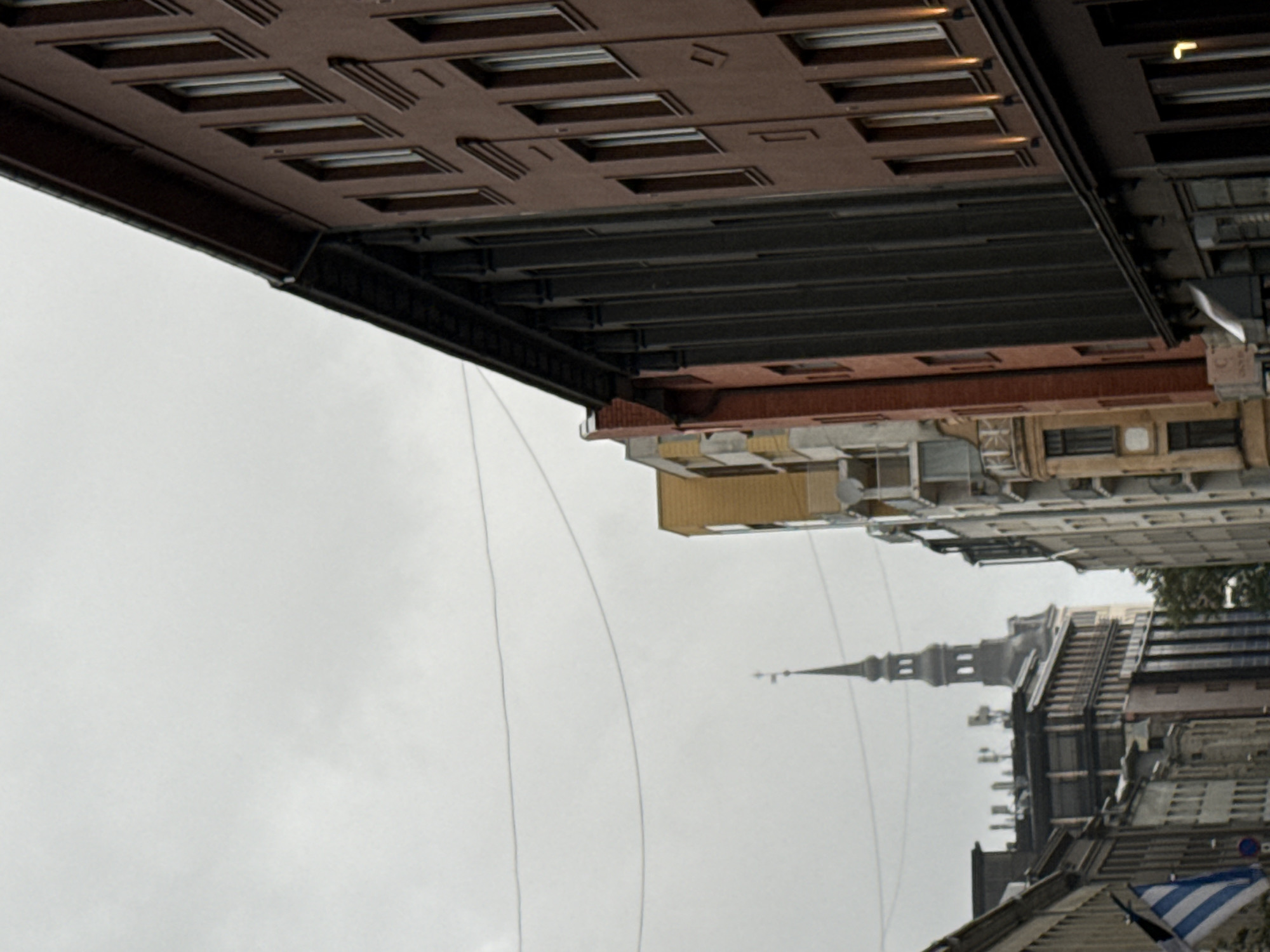 This is a street-level view in Tallinn, Estonia, looking down a city street with buildings lining both sides. The sky is overcast and grey. In the distance, a tall, pointed church spire with multiple tiers is visible above the rooftops. The buildings vary in architectural style and color, with some appearing older and more ornate, while others are more modern. Some of the buildings have multiple stories with visible windows. There are also power lines stretching across the sky. On the left side of the image, a flag with blue and white stripes is visible. The scene depicts a typical urban environment on a cloudy day.