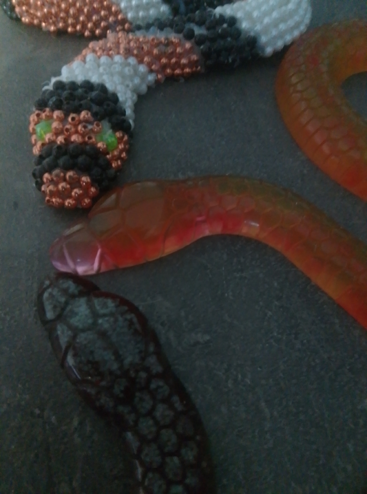 The image shows a beaded snake sculpture and three gummy worms on a dark, textured surface. The beaded snake has a head with green eyes made of small beads, and its body is constructed from white, black, and copper-colored beads. It appears to be coiled. To the right of the beaded snake are two translucent gummy worms. One is reddish-orange and the other is darker, appearing black or dark gray, with a hexagonal scale pattern. The background is a dark, uniform surface, possibly a table or countertop. The lighting is dim, casting soft shadows and highlighting the textures of the objects. There are no people or any discernable elements of a specific urban location.