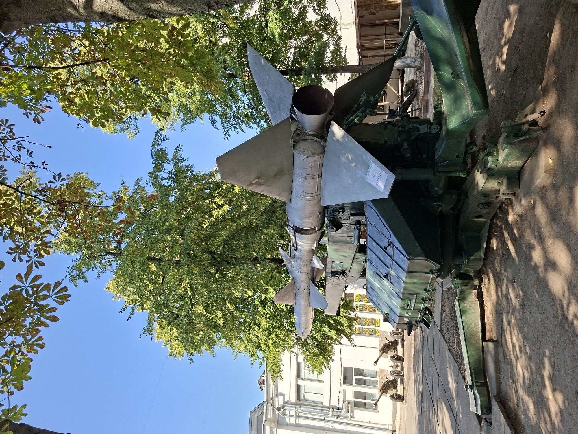 This is a medium shot, eye-level outdoor photograph of a military missile launcher in Chișinău, Moldova, likely at the National Museum of Military History. The missile is mounted on a large, dark green wheeled launcher which is positioned on a paved surface. The launcher has a complex mechanical structure with various arms and supports. The missile itself is long and silver with prominent fins at the rear. A white sign with black text reading "NU URCAȚI" (Do not climb) is visible on one of the fins. In the background, a white building with multiple windows is visible. To the left of the missile launcher, two smaller artillery cannons are positioned. The scene is framed by large trees with green and some autumnal-colored leaves, casting shadows on the ground. The sky is clear and blue. There are no people visible in the image. The lighting suggests it is a sunny day.