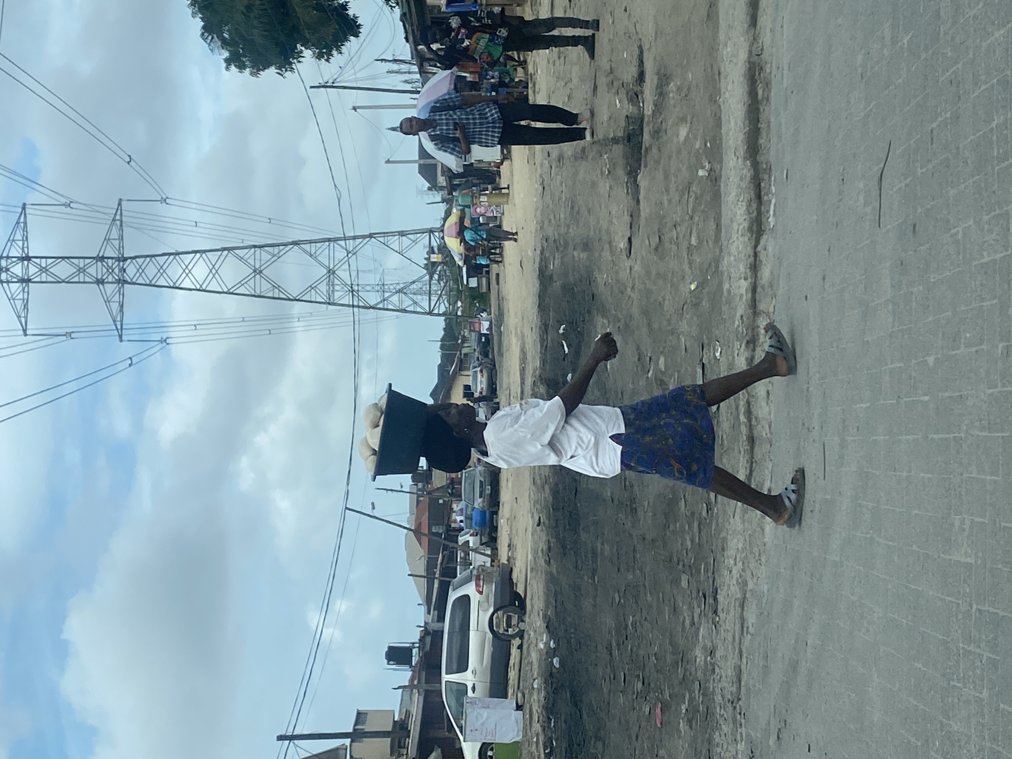 The image depicts an outdoor scene, likely in a semi-urban or rural area. A woman is walking from left to right across the frame, carrying a dark-colored bucket filled with spherical, light-colored objects on her head. She is wearing a white t-shirt and a patterned blue and white skirt. Her feet are clad in light-colored sandals.
In the background, there are several structures, including buildings with corrugated roofs and what appear to be market stalls or shops. A silver minivan is parked on the left side of the image. Power lines and a tall, lattice-like electrical tower dominate the upper portion of the frame. Other people are visible in the mid-ground, some under colorful umbrellas, and there are parked vehicles. The ground is a mix of dirt and paved areas, with some puddles or patches of dark, wet earth. The sky is overcast with visible clouds.