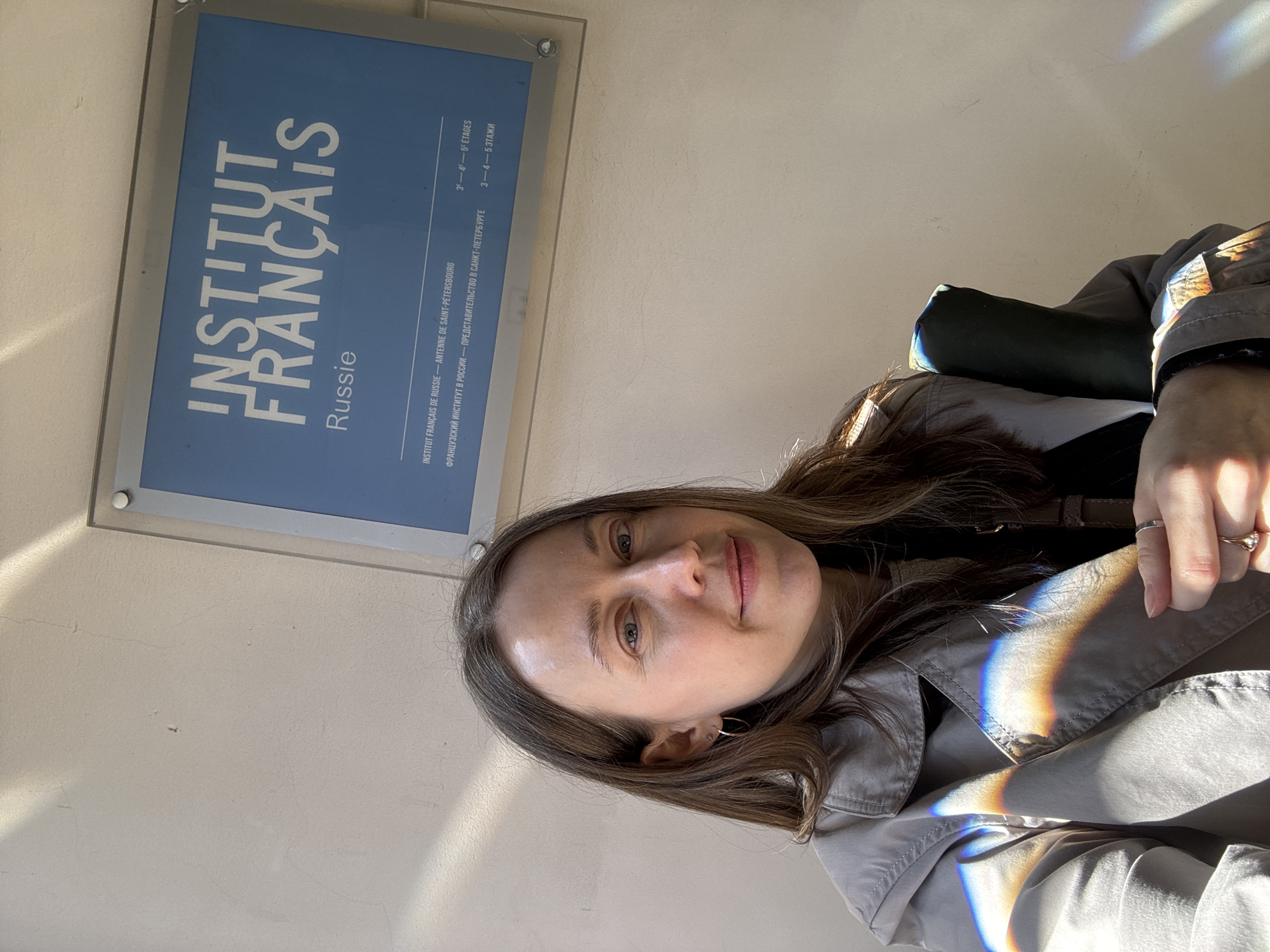 The image depicts a woman in a gray trench coat taking a selfie in front of the Institut Français in Saint Petersburg, Russia. The sign indicates the building houses the French Institute in Russia, specifically the Saint Petersburg branch, and notes that the institute occupies the 3rd, 4th and 5th floors. Light refracts prismatically on the right side of the image, reflecting on the trench coat and woman's hand.