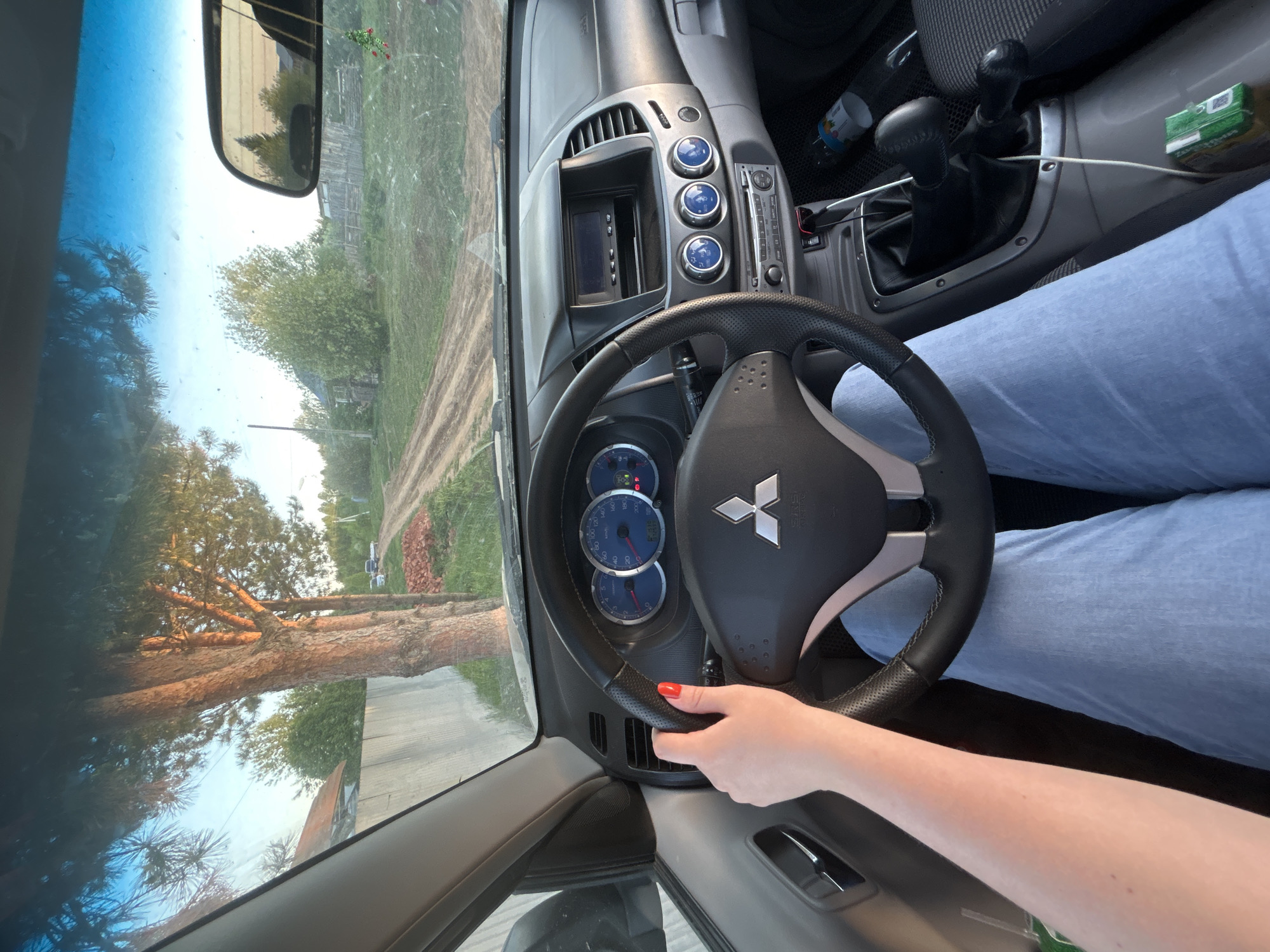 A view from inside a Mitsubishi Coltius truck, capturing the dashboard, steering wheel, and part of the driver. The driver's right hand rests on the steering wheel, adorned with red nail polish. The truck's interior includes a manual transmission, dashboard gauges, and a radio console. The view through the windshield shows an outdoor scene, suggesting a rural environment outside Фоки, Russia. A large tree and a dirt road are visible, providing context for the location.