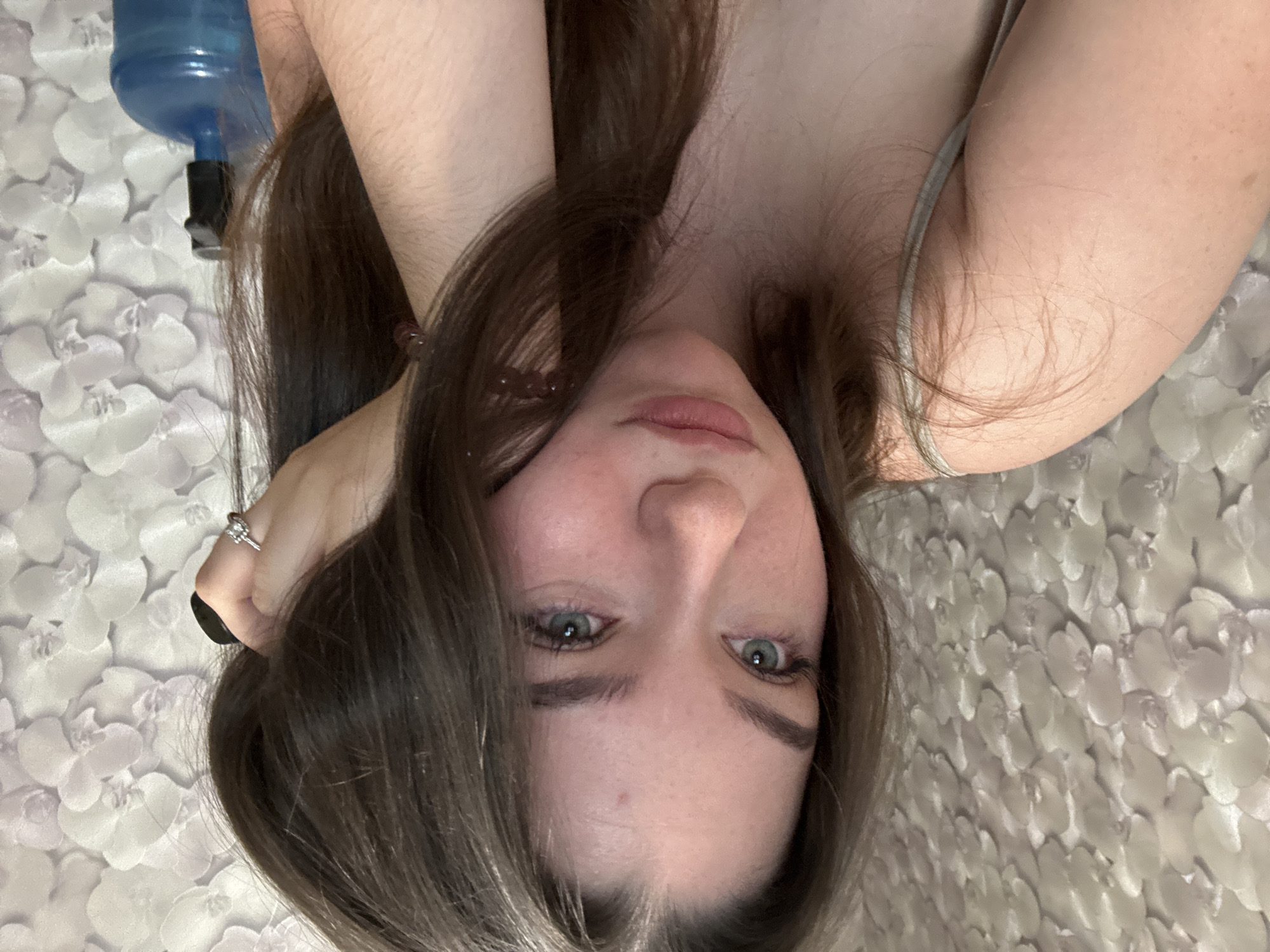 A young woman with long, dark brown hair and blue eyes is positioned in the center of the frame, looking slightly to her left with a pensive expression. Her left arm is resting on her cheek, and she is wearing a thin, light-colored strap top. She has a ring on her left hand. In the background, a floral patterned wallpaper is visible. To the right of the woman, a blue water cooler bottle with a black spout is present. The image appears to be taken indoors, likely in a residential setting within Zarechnaya (Gamovskoe S/P), Russia.