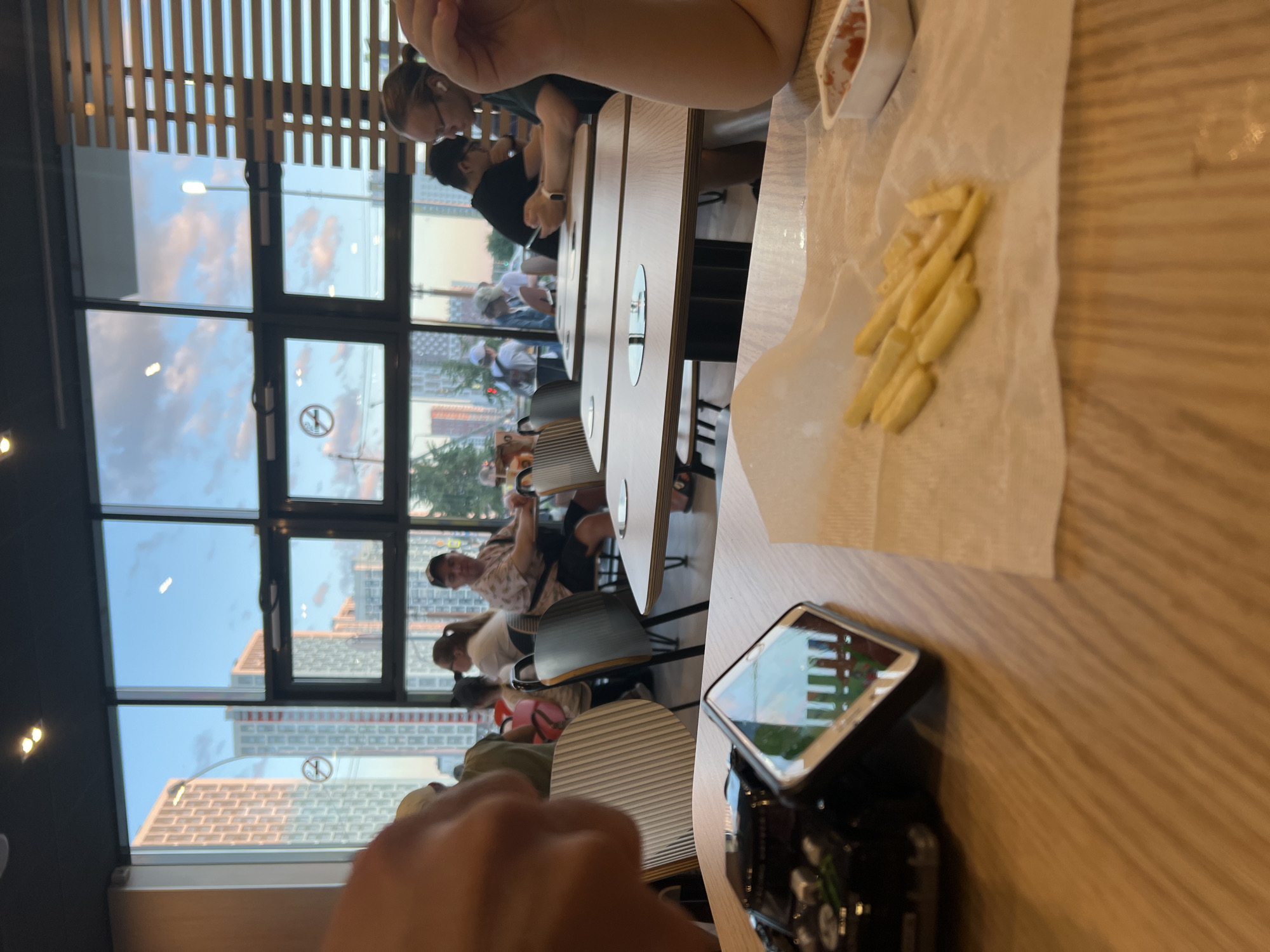 This media file depicts an indoor scene at a restaurant, likely a fast-food establishment, given the presence of French fries on a paper wrapper and a small container of sauce. The setting is a modern dining area with wooden tables and chairs, and large windows that offer a view of an urban landscape. The time of day appears to be late afternoon or early evening, as the sky visible through the windows is a soft blue with hints of pink, suggesting either sunrise or sunset. The weather outside seems to be clear.
Several people are present, seated at tables and interacting with each other. Some are looking down at their phones, while others appear to be engaged in conversation. In the foreground, a hand is visible, suggesting the photo was taken from the perspective of a diner. On the table, a smartphone is placed, its screen displaying what looks like a game or application with a grid of colored squares. A pile of golden French fries is laid out on a white paper napkin.
The view through the windows reveals tall buildings with modern architecture, characteristic of a city. The reflections on the glass indicate an indoor environment with lighting fixtures. There are no visible signs with text in the image, and the emotions of the people are not clearly discernible due to the angle and focus. The overall atmosphere seems casual and relaxed, consistent with a casual dining experience. The context of Yekaterinburg, Russia, provides a geographical reference for the urban scenery.