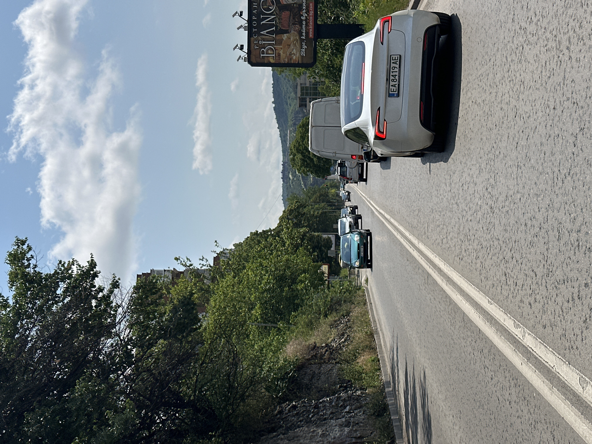 The image captures a bright, sunny daytime scene on a road near Veliko Tarnovo, Bulgaria. The perspective is from a vehicle, looking forward at a line of traffic.
The most prominent subject is the rear of a white Tesla car, which is either stopped or moving slowly. Its Bulgarian license plate, "EA 8419 AE," is clearly visible, with 'EA' indicating the region of Veliko Tarnovo. Behind the Tesla, a white van and several other cars are visible in a queue. The road surface is asphalt, marked with faded double white lines.
To the left, lush green trees and bushes dominate the foreground and midground, suggesting a natural, hilly landscape. In the distance, more verdant hills rise under a clear blue sky, dotted with scattered white clouds.
On the right side of the road, a large billboard stands tall, displaying advertising. Visible text on the billboard includes "РЕСТОРАНТ" (Restaurant), "BIANCO," and the slogan "Вкус, който помниш" (Taste that you remember), along with the website "www.bianco-bg.com". The presence of Cyrillic text and the Bulgarian license plate firmly place the scene in Bulgaria. The strong sunlight creates clear shadows, indicating a bright and clear day. There are no people visibly engaged in specific activities, but the queued vehicles suggest typical road traffic.