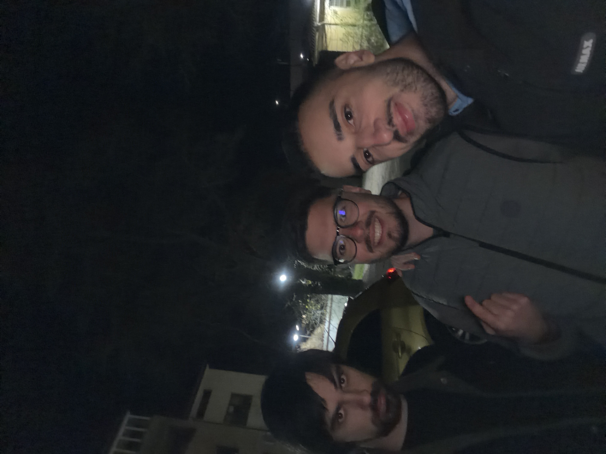 Three men are in a close, shoulder-to-shoulder shot. The man at the top is sticking his tongue out. The man in the middle wears glasses. The man at the bottom has long, dark hair. The background is dark with a building and lights visible, potentially placing them outdoors in Plovdiv, Bulgaria at night. A portion of a yellow vehicle is visible behind the man in the middle.