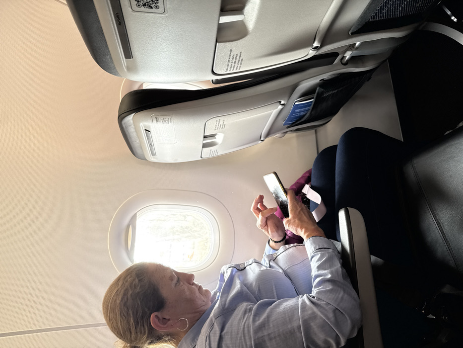 The scene is set within a commercial aircraft cabin, presumably in transit to or from the Area Metropolitana Centro Occidente (AMCO), Colombia. The specific ground location within AMCO is not discernible.
An adult female, appearing middle-aged, with light brown hair pulled back, occupies a window seat on the aircraft's left side. She wears a light blue collared button-up shirt, dark trousers, a silver hoop earring in her left ear, and a dark beaded bracelet on her left wrist. Her gaze is directed downwards at a dark-colored smartphone, which she holds horizontally with both hands, actively interacting with its touchscreen with her left index finger. A purple or magenta fabric item is draped across her lap, partially covering a fastened seatbelt.
To her right, the seat directly in front, an unoccupied passenger seat, features a black upholstered back and a light gray plastic shell. This shell includes a fold-down tray table mechanism, a safety card holder, and a mesh pocket containing blue-covered documents. A QR code and the "Avianca" brand name are visible on the upper portion of this seat-back. The aircraft interior exhibits light beige overhead panels and a circular window adjacent to the seated female, through which bright ambient light and indistinct exterior features are visible. The flooring is dark-colored.