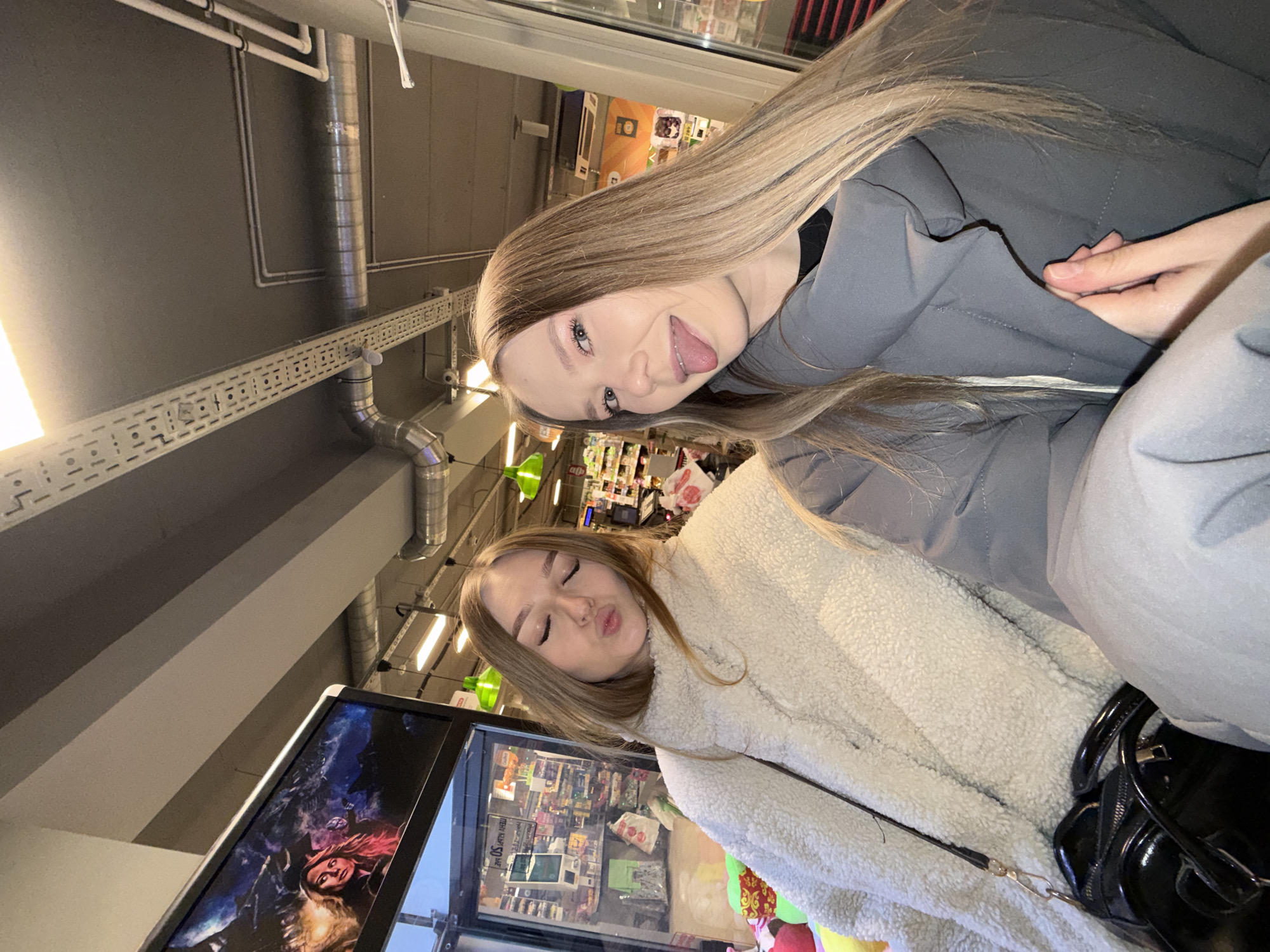 Two young women are featured in a playful indoor photo, likely a selfie, taken at an angle within a commercial setting.
The woman positioned higher in the frame has long, straight blonde hair and is wearing a grey jacket. She has a wide, playful smile, sticking her tongue out towards the camera. Her eyes are light-colored.
Below her, the second woman, also with long blonde hair, wears a fluffy, cream-colored jacket. She is making a kissing face (pouting) with her eyes closed or mostly closed. A black handbag with a shoulder strap is visible near her.
The background indicates an indoor commercial space, possibly a store or supermarket. The ceiling features an industrial design with exposed silver ventilation ducts, white pipes, and cable trays. Fluorescent lighting fixtures illuminate the area. In the mid-ground, a display screen shows an image of a person, possibly a woman with reddish hair, against a dark, dramatic backdrop. Further behind, brightly lit shelves stocked with various products are visible. Some blurry text on price tags includes "РУБ" (Ruble) and numbers like "50", strongly suggesting a location in Russia, specifically Chaikovsky as per the context. The overall atmosphere of the photo is casual and lighthearted.