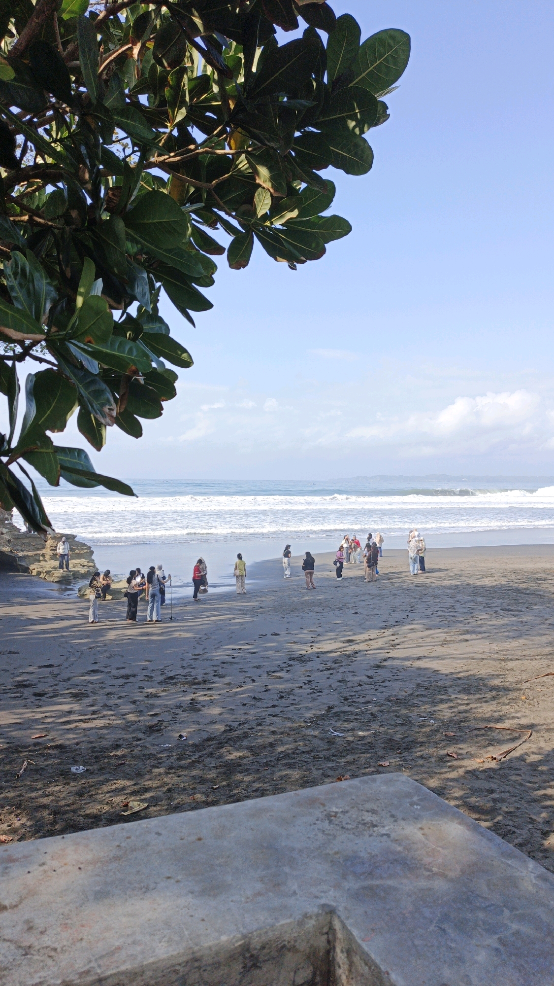 The image depicts a sunny day at Desa Ciliang beach in Indonesia, with a group of people gathered on the wet sand near the shoreline. The scene is framed by large green leaves and branches of a tree in the foreground, casting shadows on the sand. The ocean, with moderate waves and white foam, stretches towards the horizon under a clear blue sky dotted with a few wispy clouds. The people, dressed in casual attire, appear to be observing the ocean, taking photos, or conversing. Some individuals are standing, while others are sitting or squatting. A concrete structure is visible in the lower right portion of the frame, possibly a bench or a viewing platform. The overall atmosphere is relaxed and leisurely, suggesting a typical day at a popular beach destination.