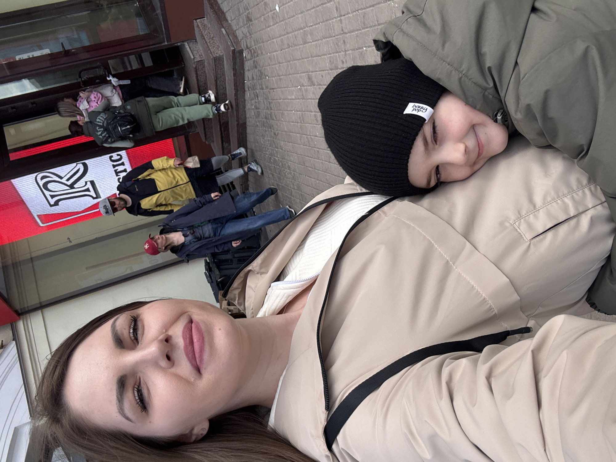 A young woman and a child are posing for a selfie outside a store in Moscow, Russia. The woman is lying down, facing the camera with her head to the left of the frame. She has brown hair, fair skin, and pink lipstick. The child is lying on top of her, with their head near her shoulder. They are wearing a black beanie with a white tag and a green jacket.
In the background, several people are walking on the sidewalk. Behind them is a storefront with a large red and white sign with the letters "R STIC" visible. The facade of the building and the sidewalk are constructed from light-colored stone or concrete.