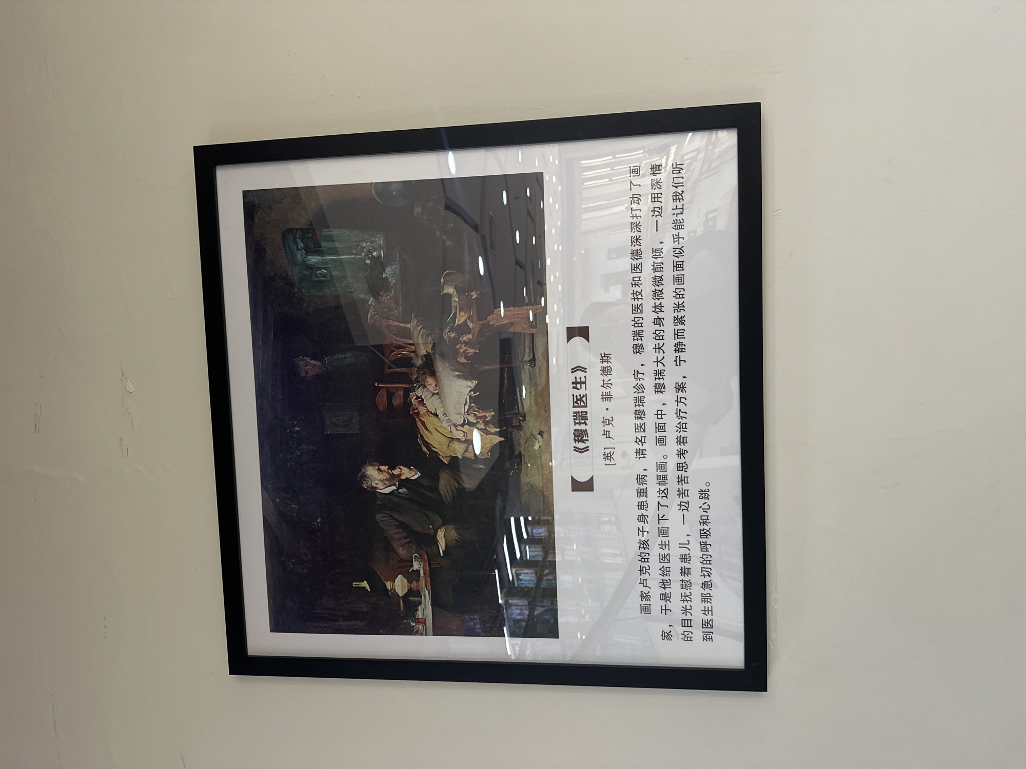 A framed picture of an oil painting hangs on a light beige wall. The painting is titled “The Doctor” (《穆瑞医生》) by Luke Fildes (卢克·菲尔德斯). The painting depicts a doctor leaning over a child lying on a bed, with medical instruments and books in the background. A lamp is on the table in front of the doctor. The Chinese text next to the painting explains that the artist’s child was seriously ill and the doctor’s skill and morals deeply moved him, so he painted this picture. The text further describes the doctor’s pose and the tension in the scene.
Based on the location metadata, the picture was taken in the city of Chongqing High-tech Zone, China.