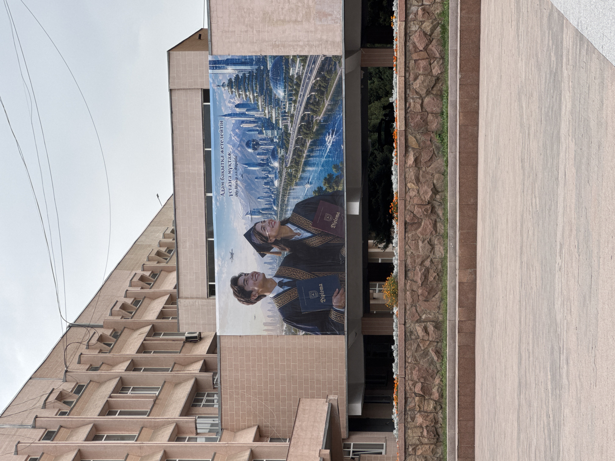 A large banner is displayed on the facade of a building in Almaty, Kazakhstan, depicting a futuristic city skyline with a river and a highway. Two young adults, a male and a female, both wearing graduation gowns and caps, stand in the foreground. The male is holding a blue diploma, and the female is holding a maroon diploma. Both are smiling and looking towards the right. The building behind them has a distinctive architectural style with a pale pink facade and repeating window patterns. In front of the building, there is a stone wall with flowers and a paved plaza. The sky is overcast.