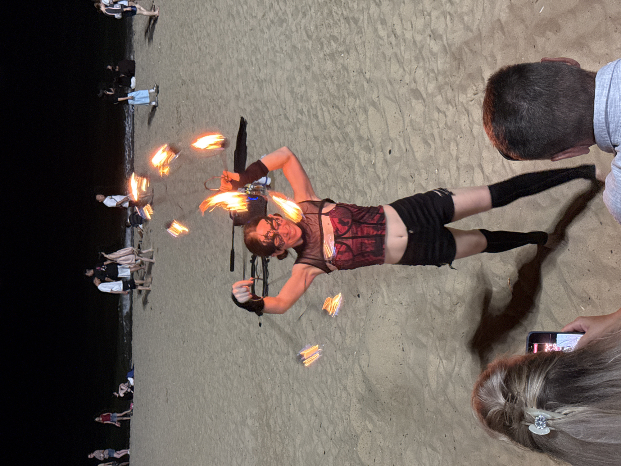 At night, on a sandy beach in Nha Trang, Vietnam, a performer in a red and black corset, black shorts, and black thigh-high socks is spinning fire. The performer, who appears to be female, is wearing decorative facial makeup and is smiling. The fire is creating bright, blurred streaks of light in the air around her. Several other people are visible in the background, some sitting and some standing, further down the beach and near the water. In the foreground, the back of a woman's head with a clip in her hair and a person's head are visible, both appearing to be watching the performance. The sand is the dominant texture of the environment.