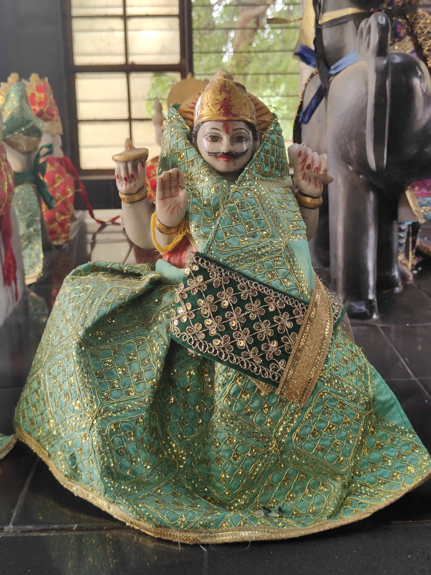 The image shows a statue of a deity, possibly Hindu, adorned with a green and gold intricate fabric garment. The statue has a painted face with a mustache and is wearing a golden crown. Its hands are raised, with one in a gesture of blessing or protection. The statue is seated, and the fabric drapes around it, creating a voluminous skirt effect. In the background, a window with multiple panes is visible, suggesting an indoor setting. To the right of the statue, a large, dark, elephant-like sculpture is partially visible. The overall scene appears to be within a temple or a similar religious or cultural space. The image does not provide any specific information about its location within Gurgaon, India, or any details about people or actions taking place.