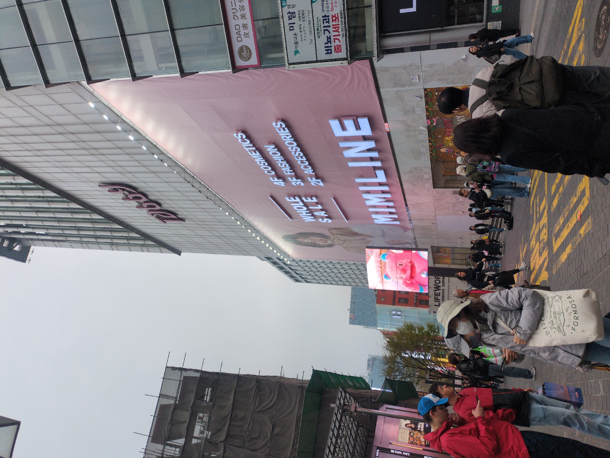 The image captures a street scene in Seoul, South Korea. A large building features a sign advertising "MIMILINE" along with "4F COSMETICS, 3F FASHION, 2F ACCESSORIES". A secondary advertisement is shown, advertising the DAO Clinic, a dermatology and aesthetic clinic. The surrounding environment contains scaffolding, other storefront advertisements, and various buildings. The people in the scene are walking on the sidewalk. They are wearing everyday clothes and masks. One individual is carrying a bag with the word "FORMOSA" printed on it. The weather appears overcast.