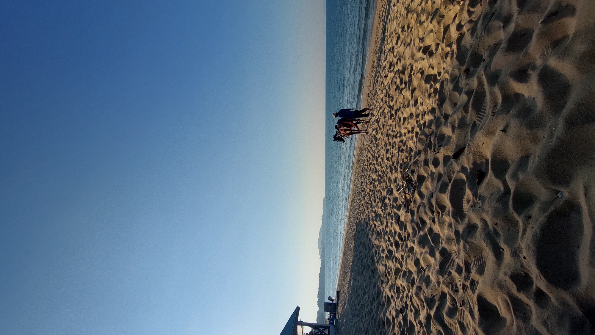 Baan Mae Nam, Thailand. A beach scene under a clear blue sky. A sandy beach occupies the right half of the frame, with varying textures and footprints visible. A person stands near the water's edge with a horse. Water is along the middle of the image. A small structure is at the bottom.